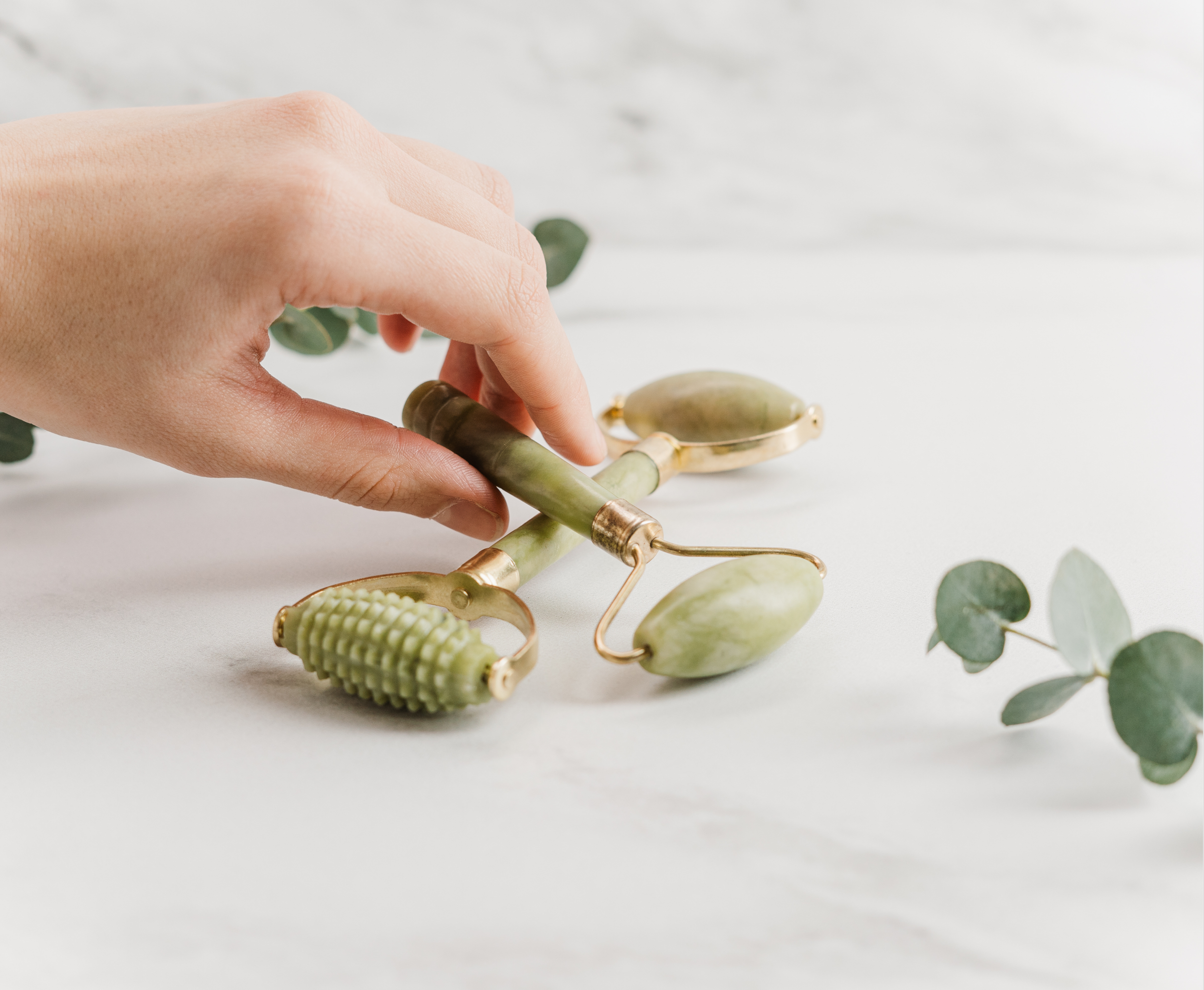 A hand holding a jade roller for facial massage, with eucalyptus leaves on a white background.