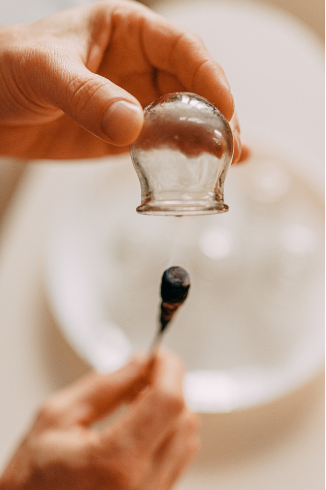 A hand holding a glass facial massage tool over a hand holding a cotton swab with some black substance, with a sink visible in the background.