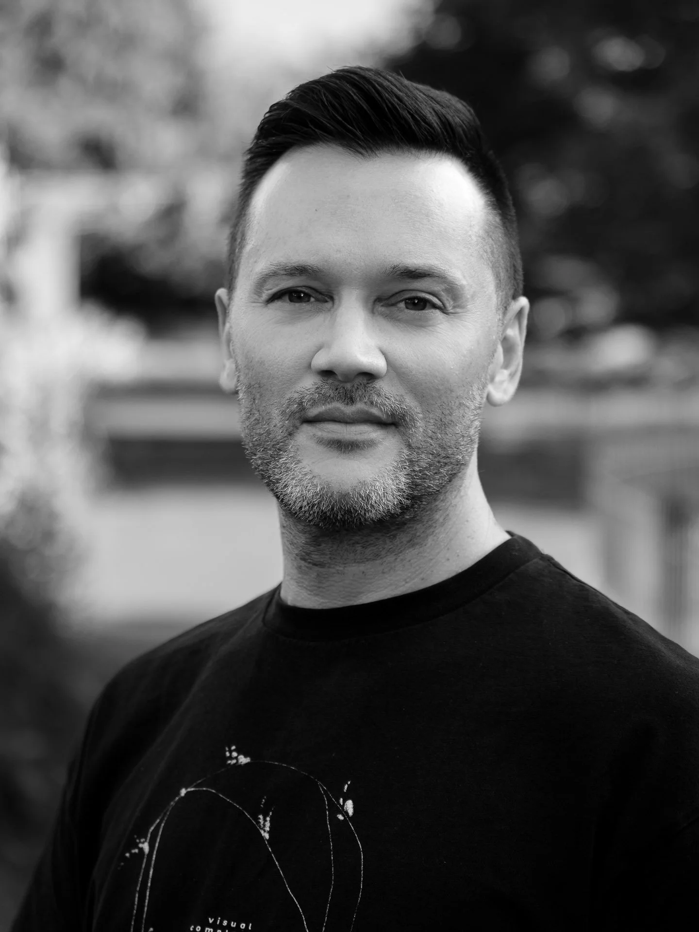 Black and white portrait of a man with short dark hair, wearing a black t-shirt, outdoors with blurred background.