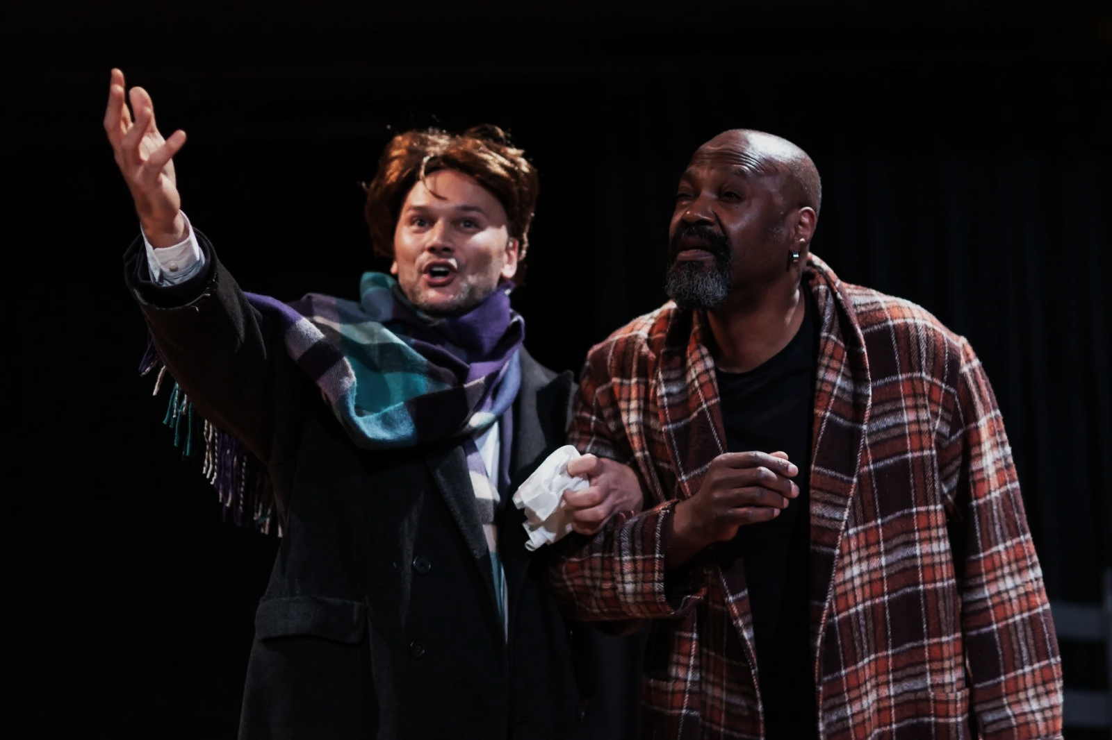 Two men are performing on stage, engaging in a theatrical scene. The man on the left, with brown hair, is gesturing animatedly with his right hand and appears to be speaking. He is wearing a black coat and a multicolored scarf. The man on the right, with a bald head and gray beard, is dressed in a red plaid jacket and is holding a white cloth in his left hand. The background is dark, emphasizing the performers.