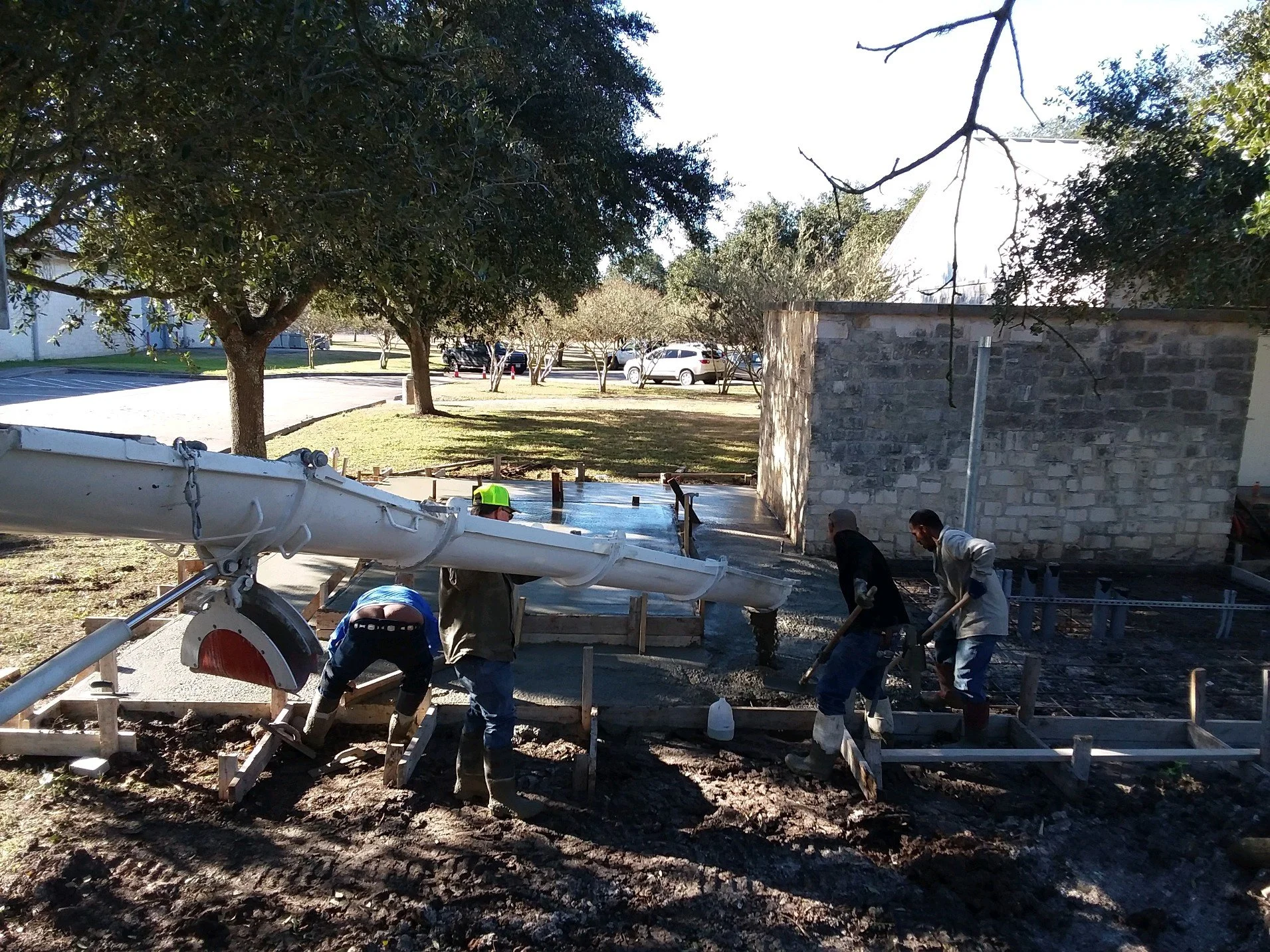 Workers pouring concrete for a sidewalk or foundation at a construction site, with trees and parked cars in the background.