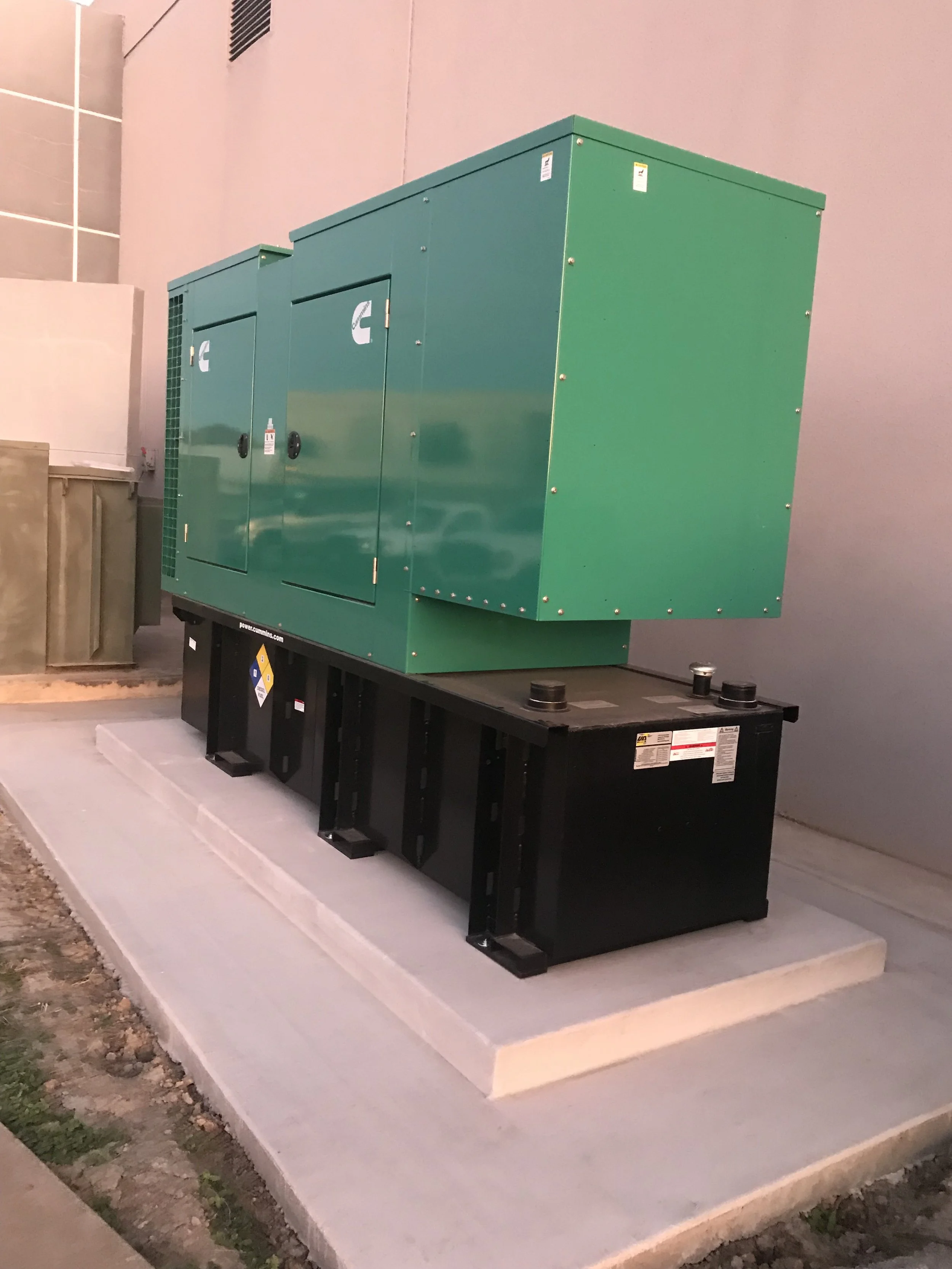 Green electrical transformer box installed on a black stand on a concrete pad, with a light pink wall in the background and a curb and dirt in the foreground.