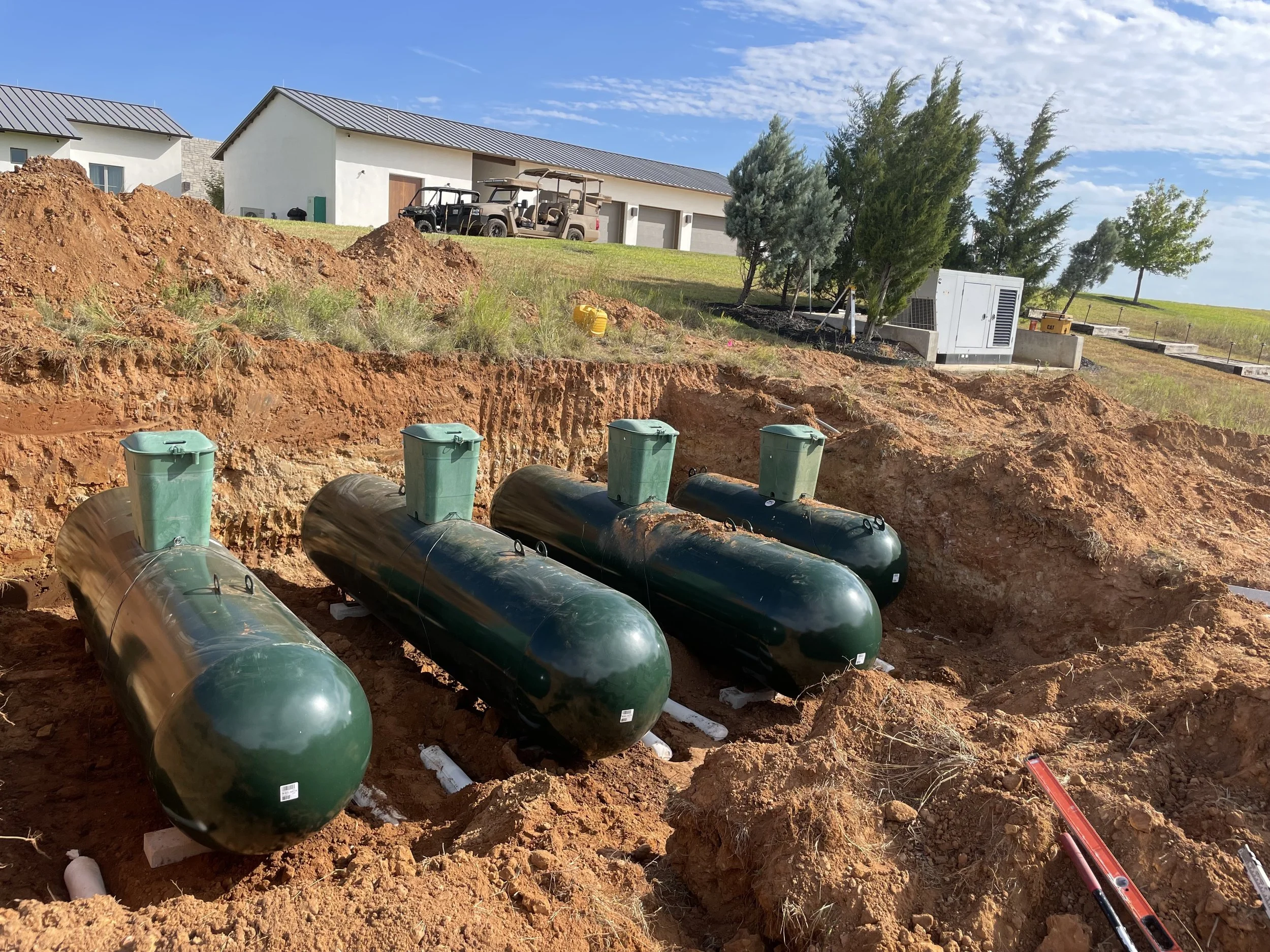 Underground pipeline installation with large green tanks, pipes, and construction equipment in front of residential houses, trees, and blue sky.