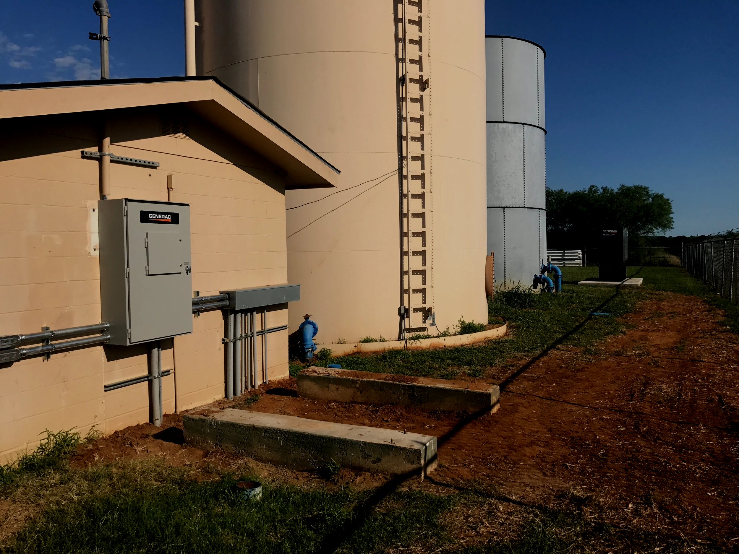 Industrial site with large beige and gray tanks, electrical equipment on the wall, electrical wiring, and blue pipes near a chain-link fence, under a clear blue sky.