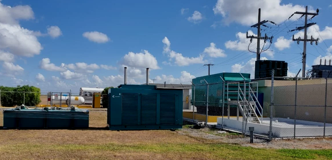 Industrial equipment on a grassy area behind a chain-link fence, including large metal boxes, a tall box with stairs, utility poles, and a partly cloudy sky.