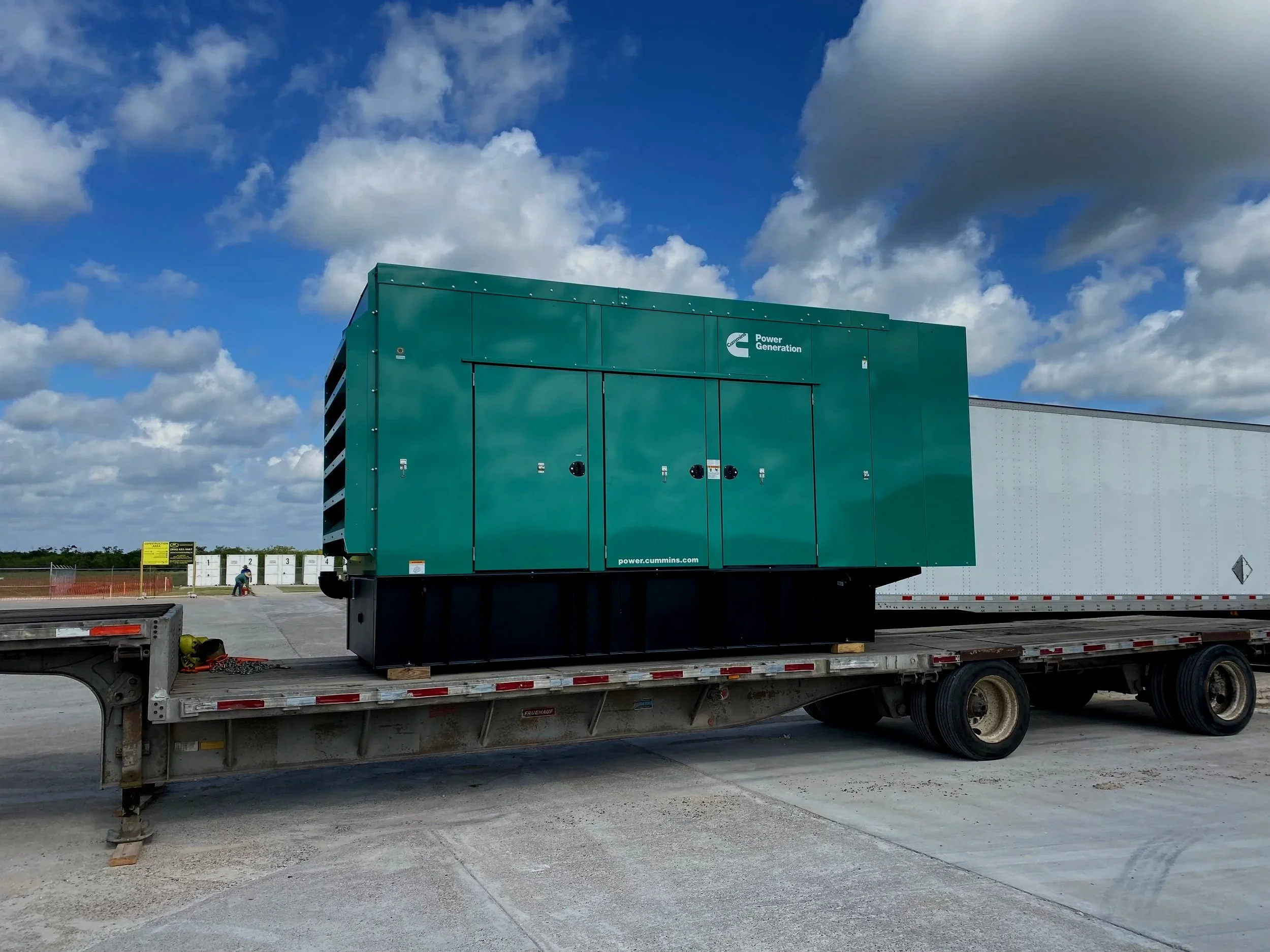 Large green industrial generator on a flatbed trailer with sky and clouds in the background.