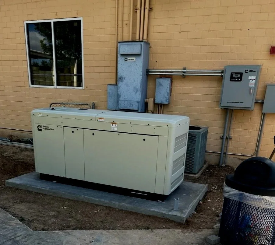 An industrial generator outside a beige brick building with a window, electrical boxes, and a trash can.