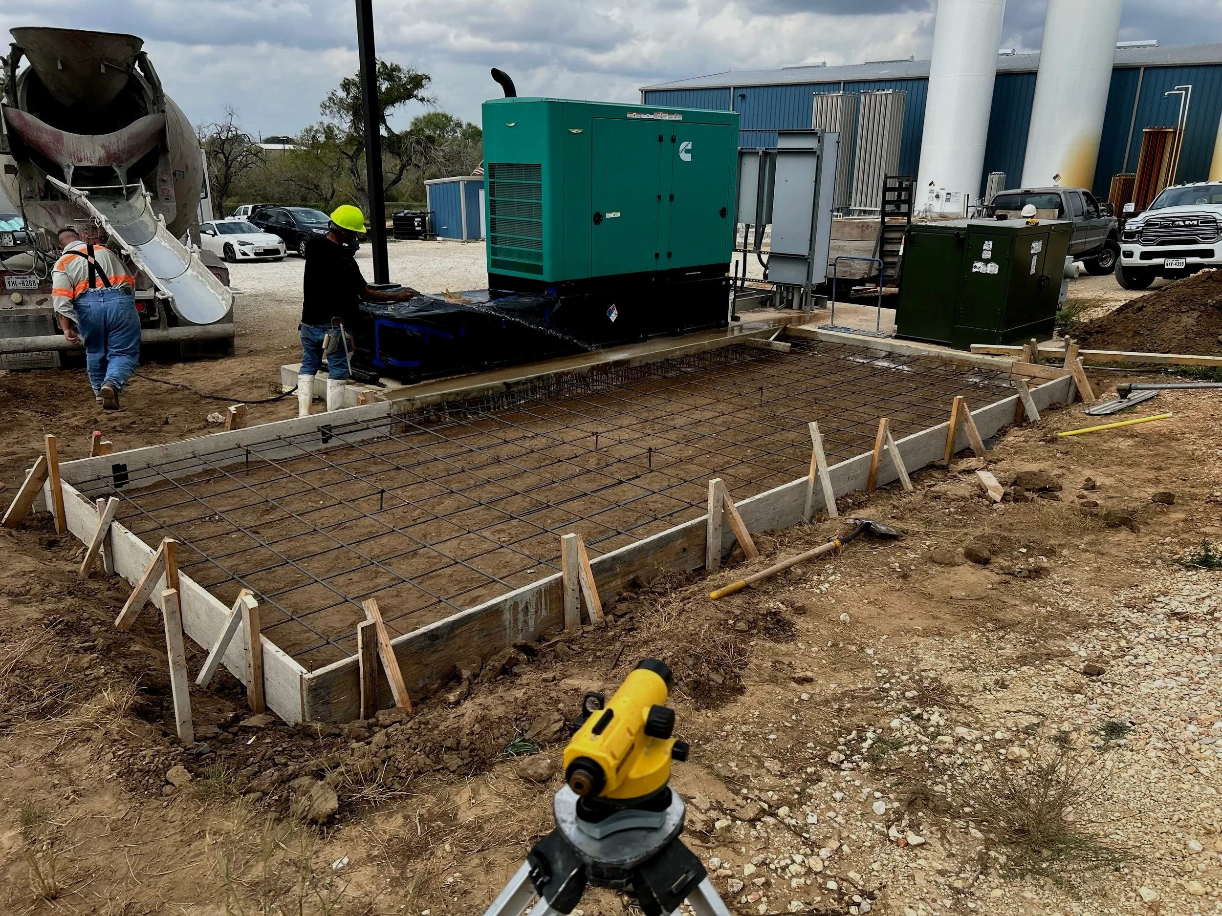 Construction site with a concrete mixer truck, workers, and equipment preparing a foundation with rebar and concrete. There are parked cars and industrial buildings in the background.