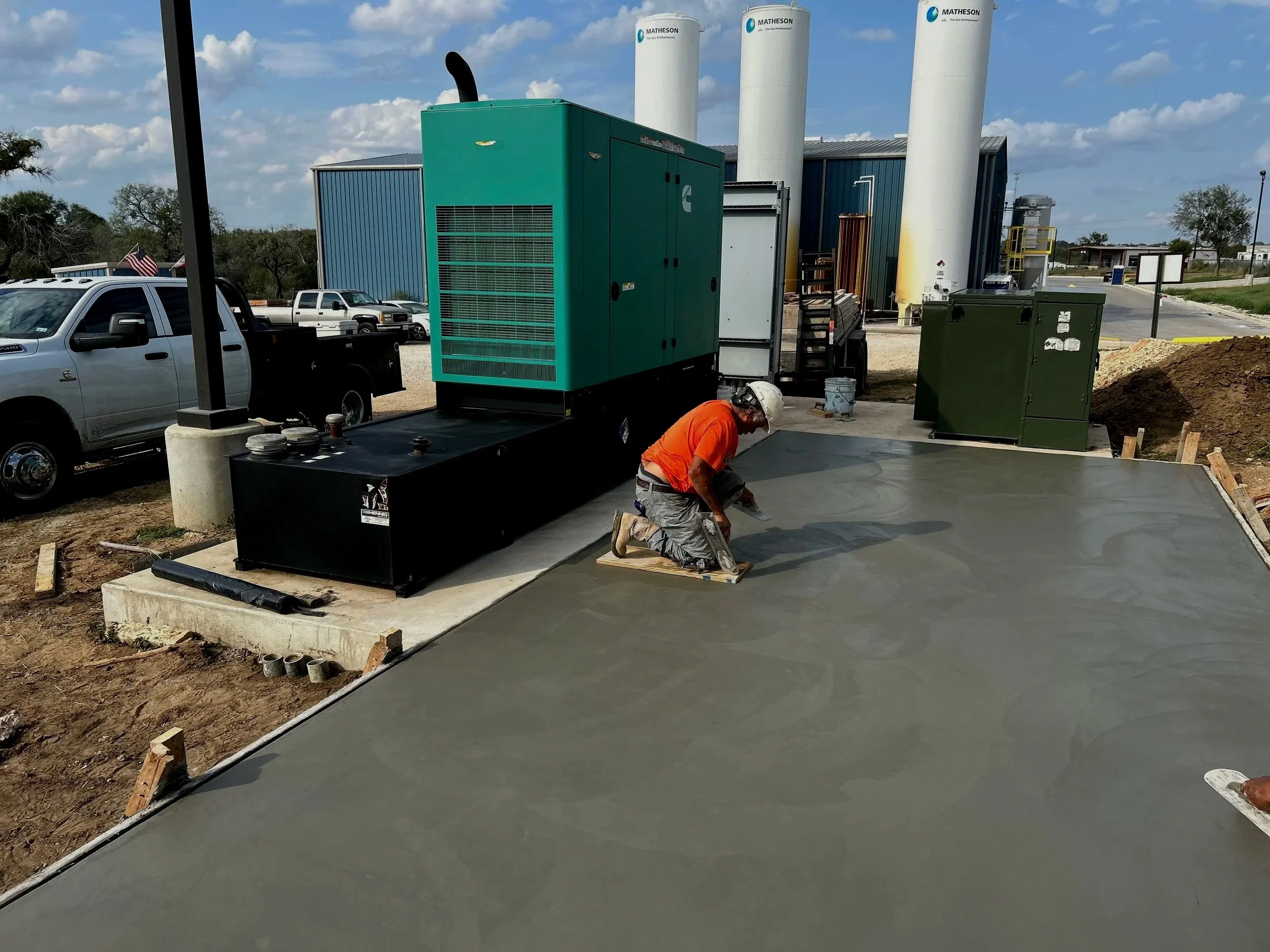 Construction worker kneeling on freshly poured concrete with construction equipment and utility vehicles nearby, and large white storage tanks in the background under a partly cloudy sky.