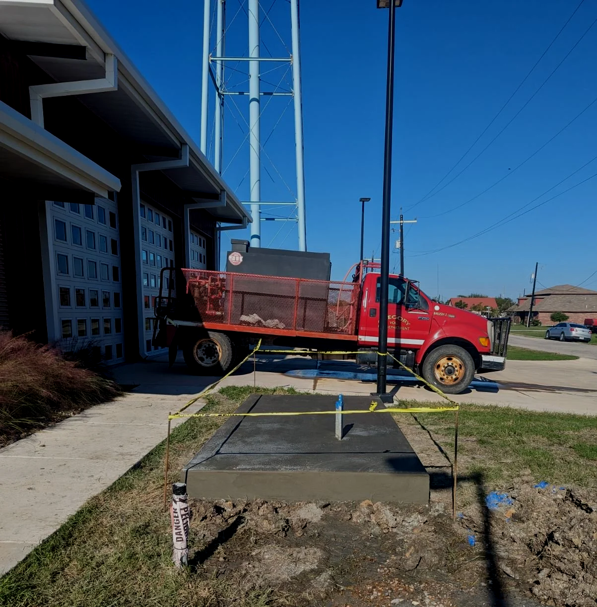 A concrete slab with exposed soil and a construction warning sign, surrounded by yellow caution tape, in front of a red utility truck parked at a construction site outside a residential building with garage doors.