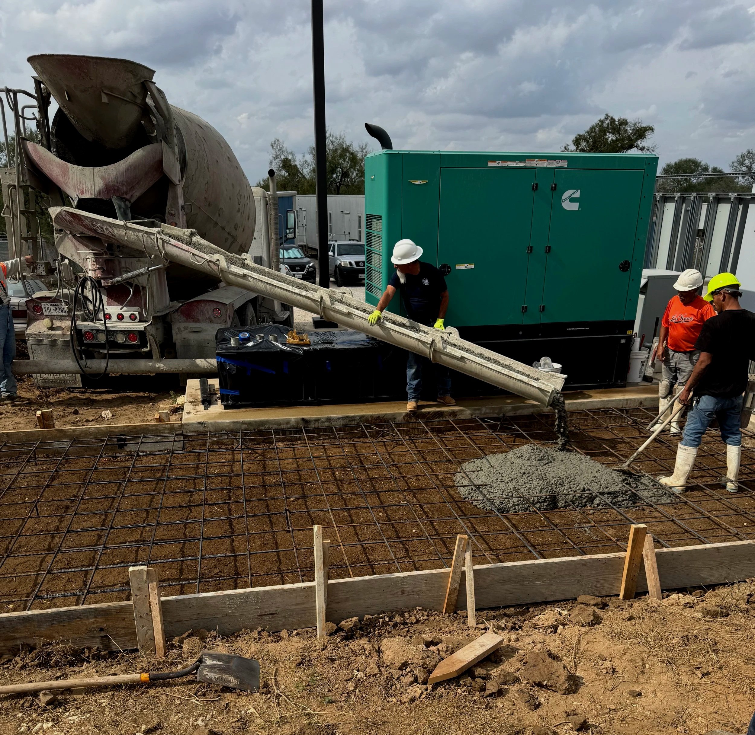 Construction site with a concrete mixer truck pouring concrete into a foundation, workers wearing helmets and safety gear, and construction equipment and materials present.