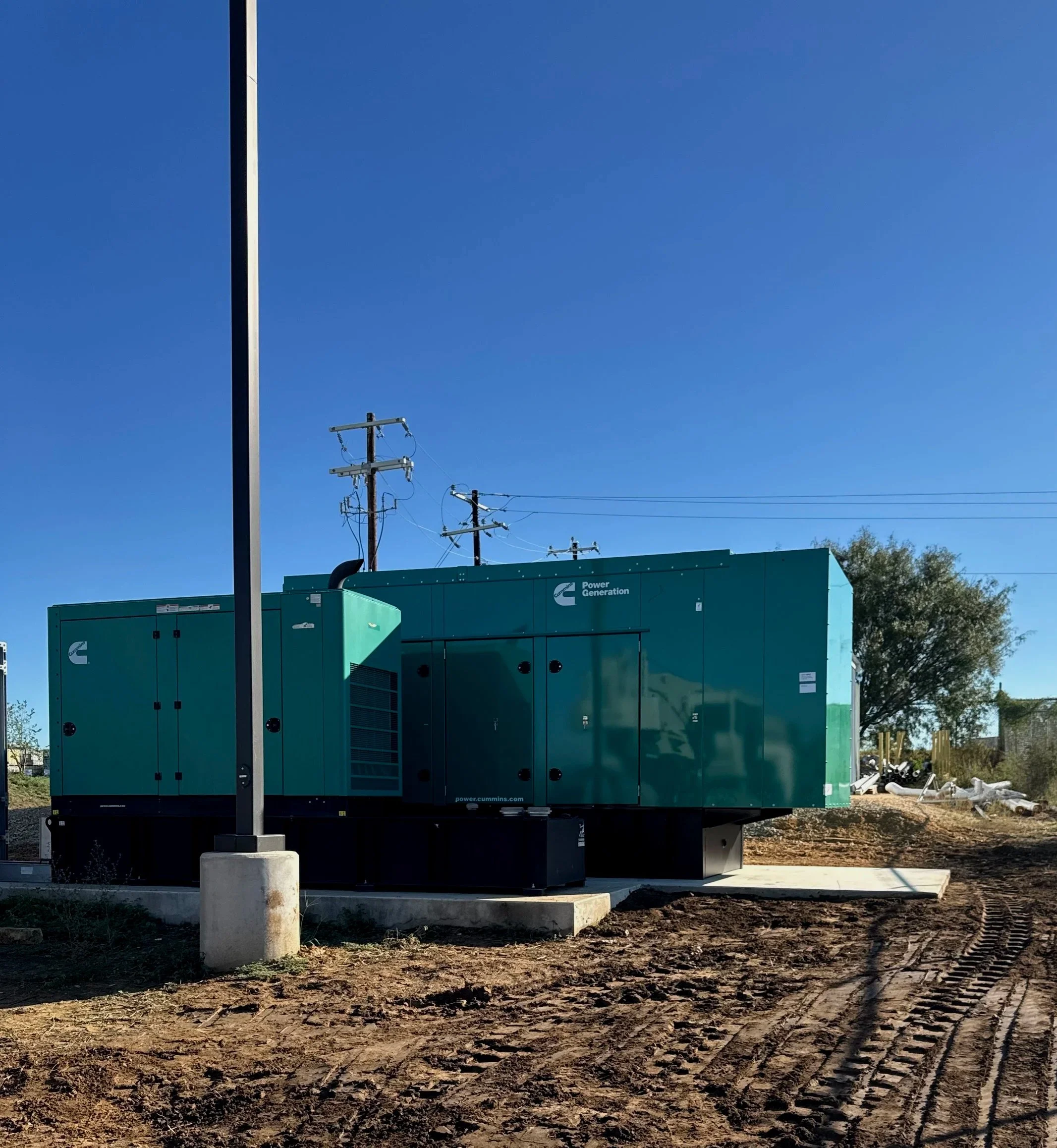 Large green industrial generator with power company logo, installed on a concrete pad, with utility poles and clear blue sky in the background.