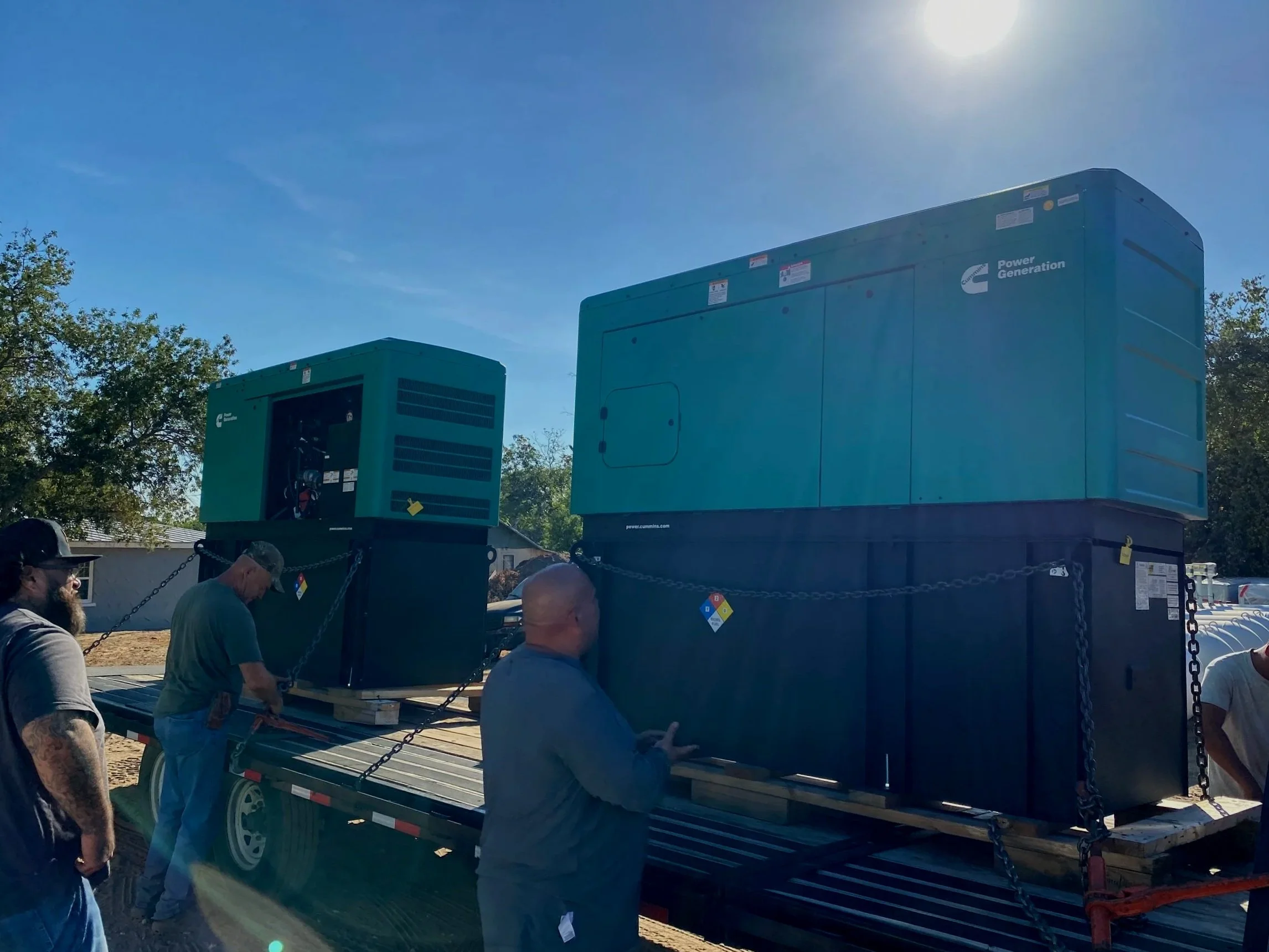 Several men are working together to unload or prepare to install large industrial equipment on a flatbed trailer outdoors beneath clear blue skies, with trees and a building in the background.