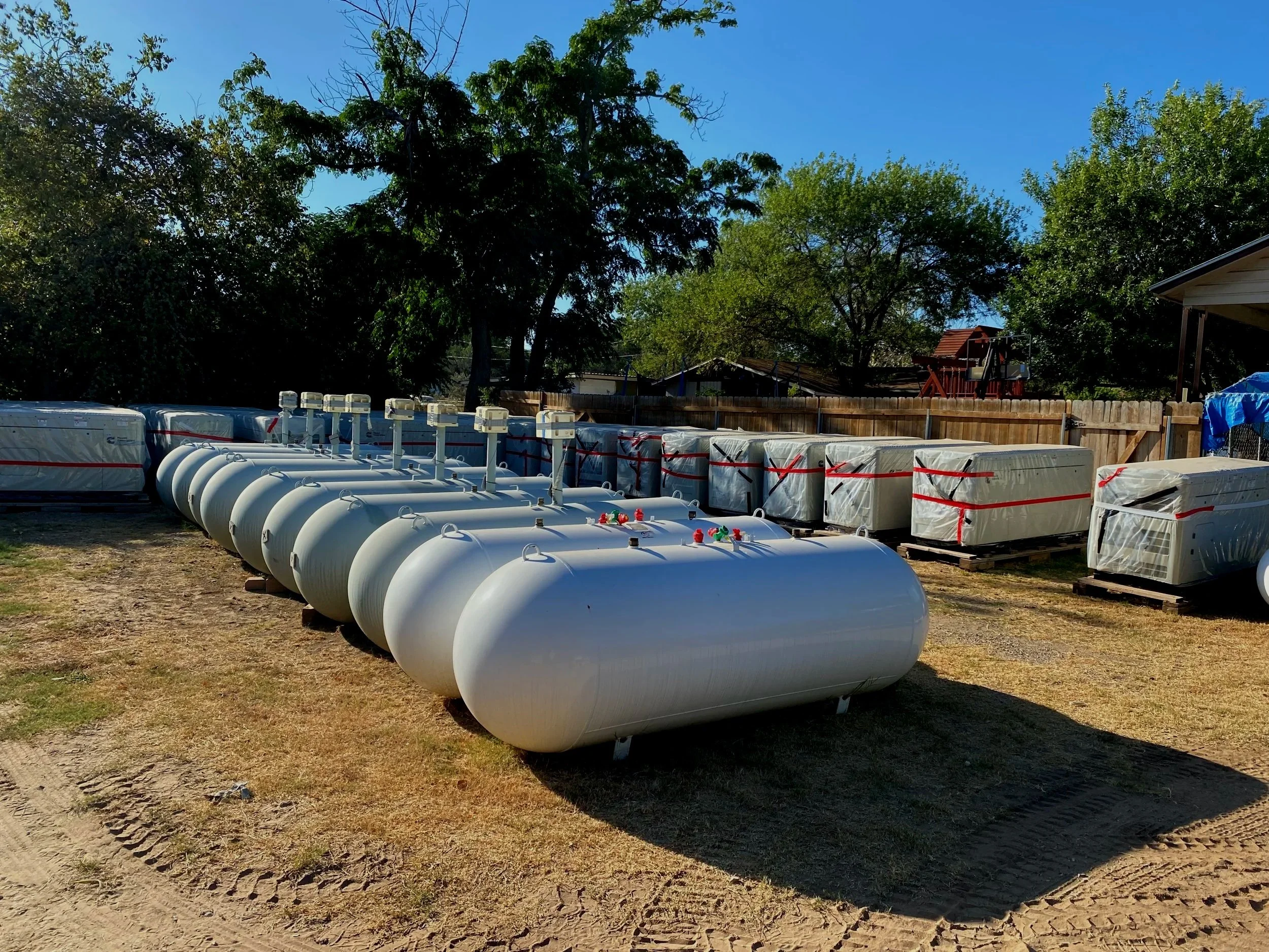 Row of large white propane tanks outdoors, surrounded by trees and a wooden fence, with blue sky overhead.