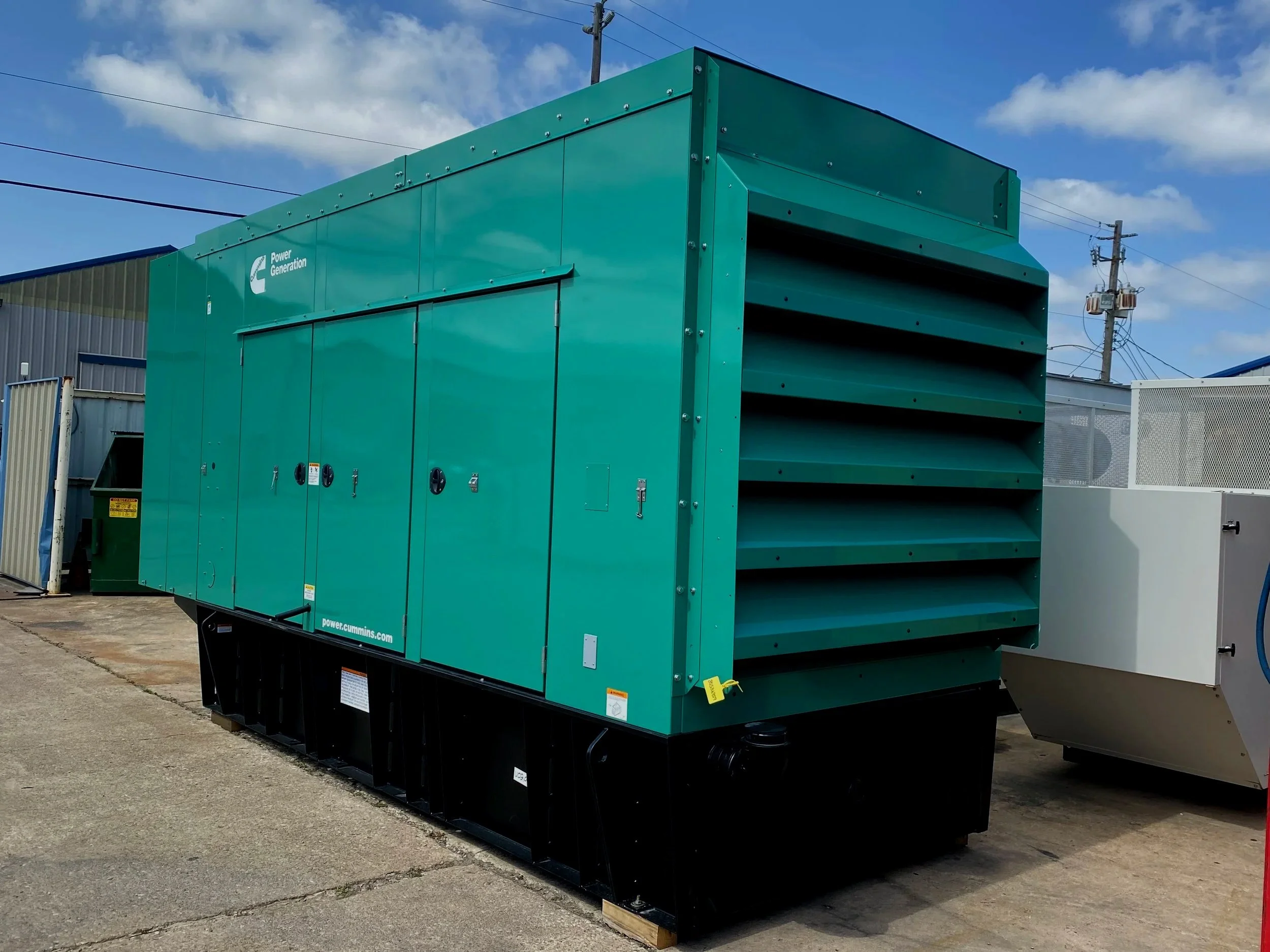A large green industrial generator outdoors on a concrete surface with blue sky and some clouds. There are utility poles and buildings in the background.
