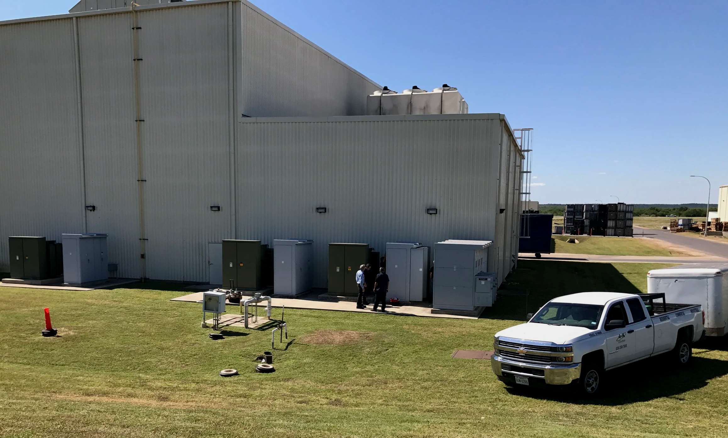 Large industrial building with metal siding and rooftop HVAC units. Several electrical boxes and utility meters are on the side. Two people are standing outside near the building, and a white pickup truck is parked in front. There are stacks of crate