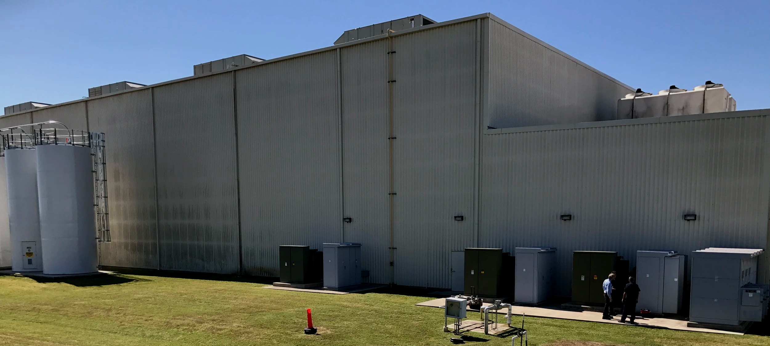 Industrial building with large metal exterior walls, cooling units on top, several utility boxes at the base, and two people standing near electrical panels.