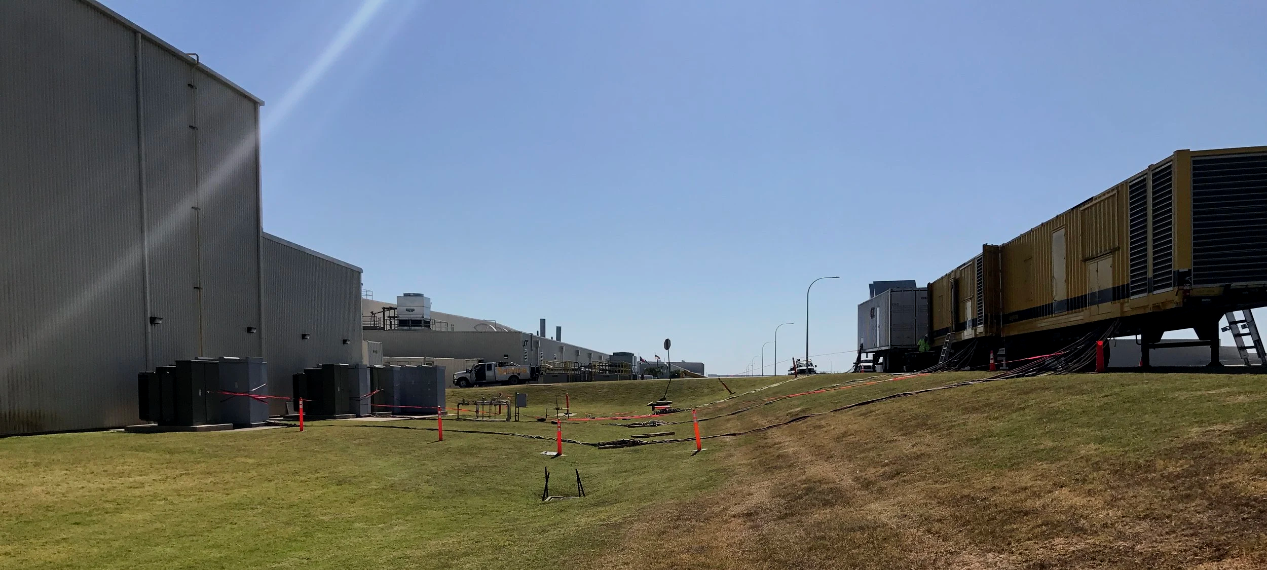 Industrial building with large HVAC units on grassy ground, trucks parked nearby, on a sunny day with clear blue sky.