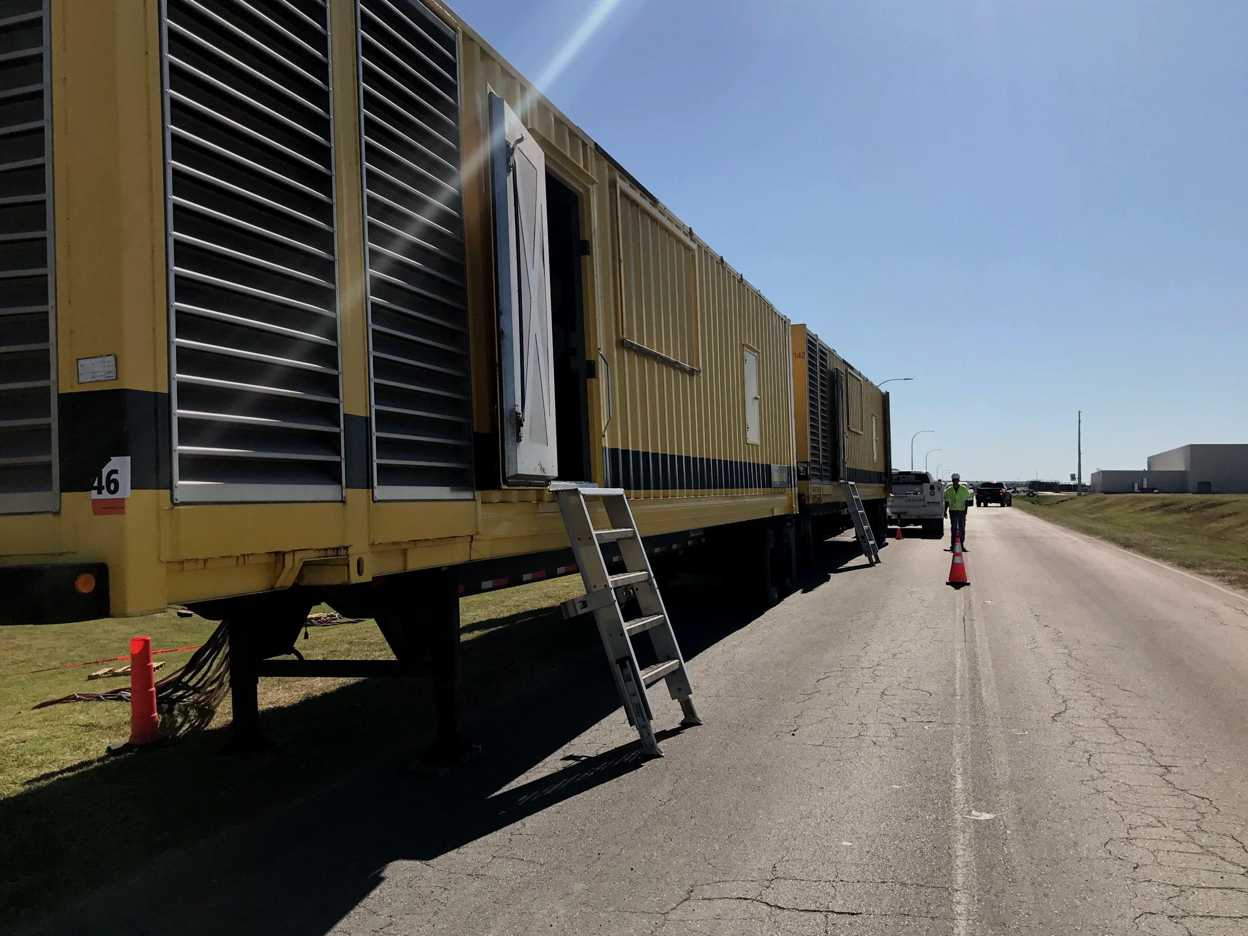 Large yellow mobile air conditioning or generator unit on a trailer, with a set of stairs leading up to an open door, set up outdoors on a paved road, with a person in safety gear walking nearby and cones marking the area, under a clear blue sky.
