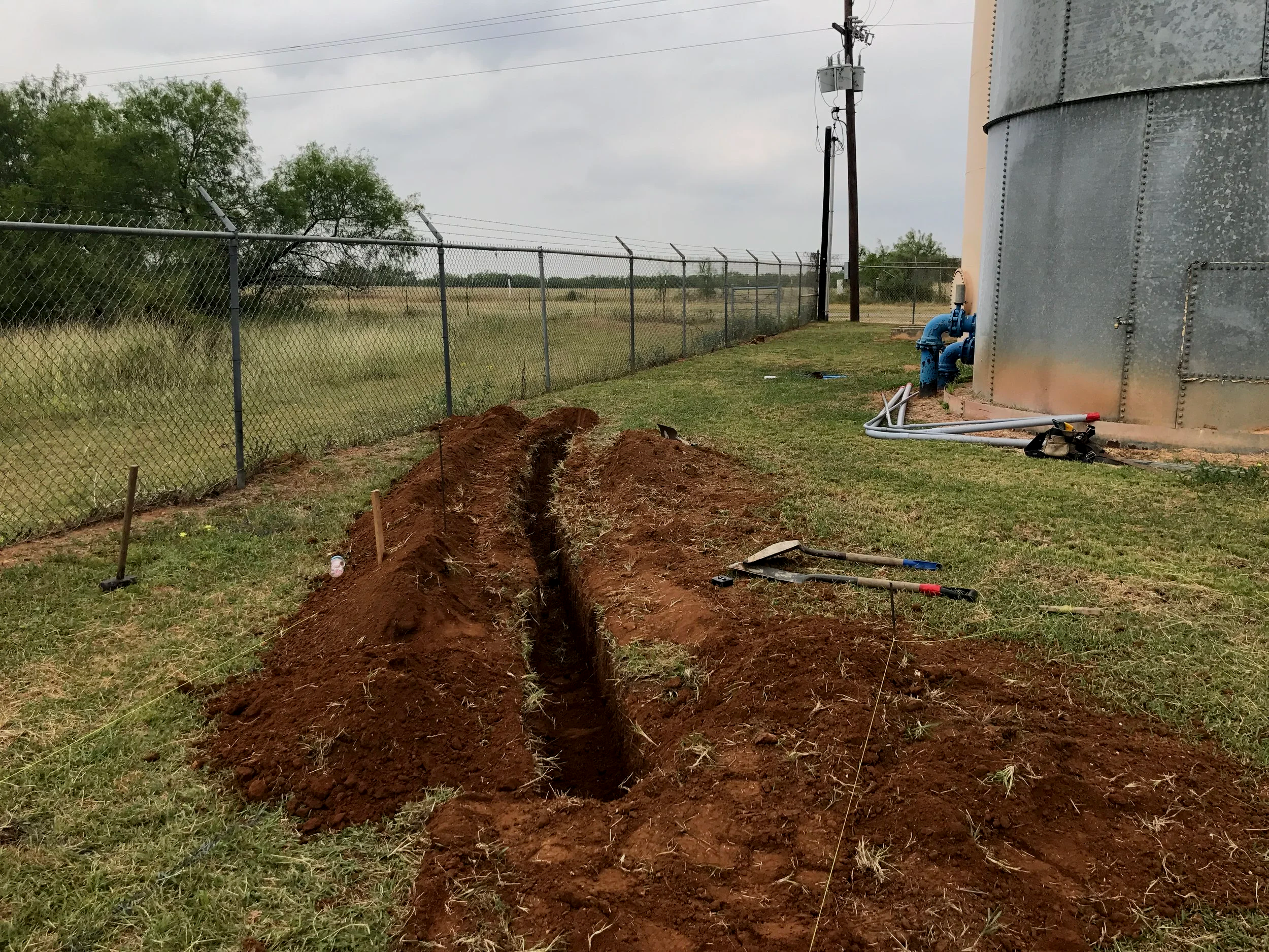 Trenched soil in an open grassy area near a chain-link fence, with trees, utility poles, and a large metallic tank in the background under a cloudy sky.