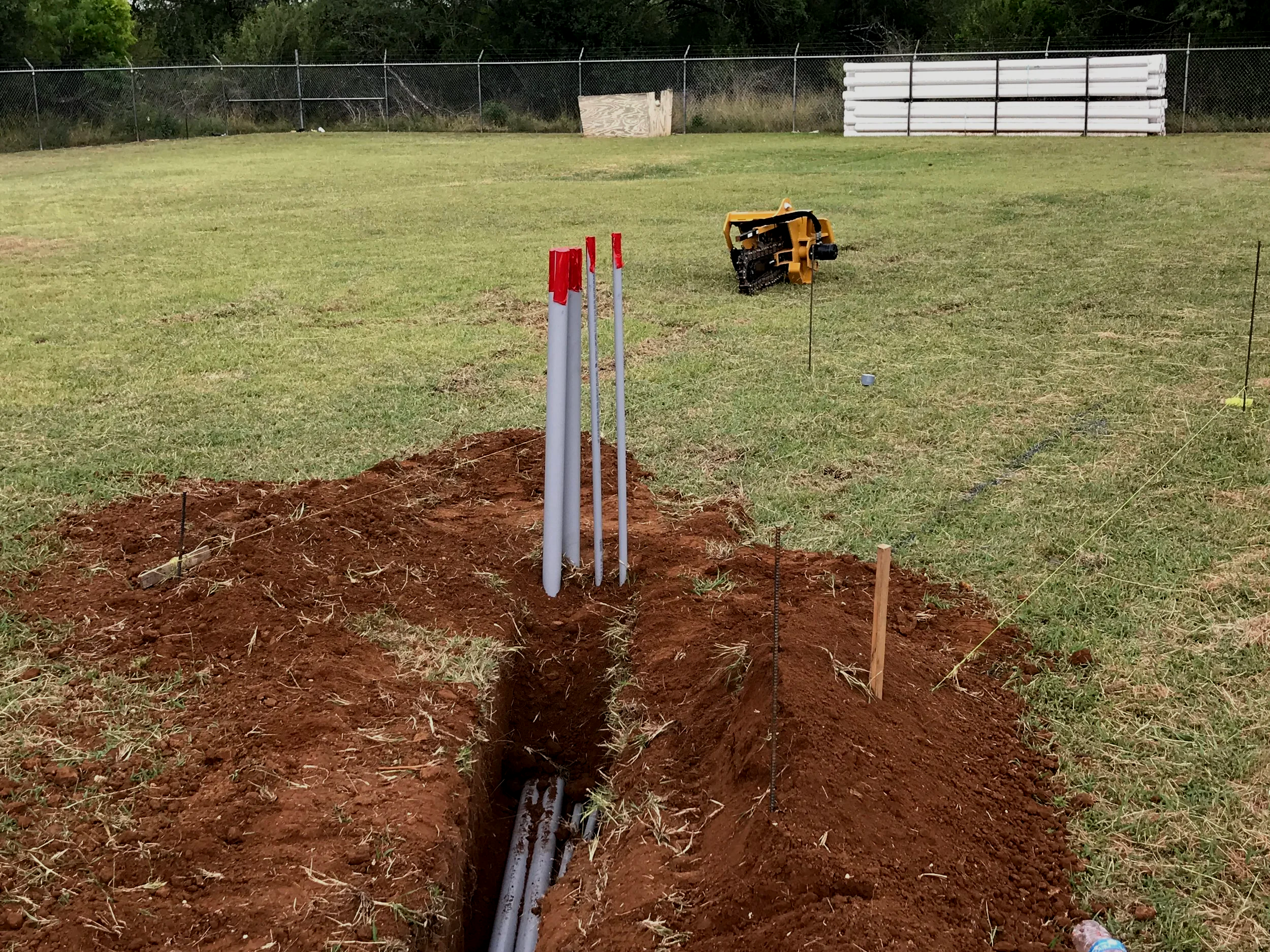 A construction site on a grassy field with a large hole in the ground, three concrete pipes in the hole, and orange-topped stakes marking the boundary. There is a yellow construction tool laying on its side, and in the background, there is a chain-li