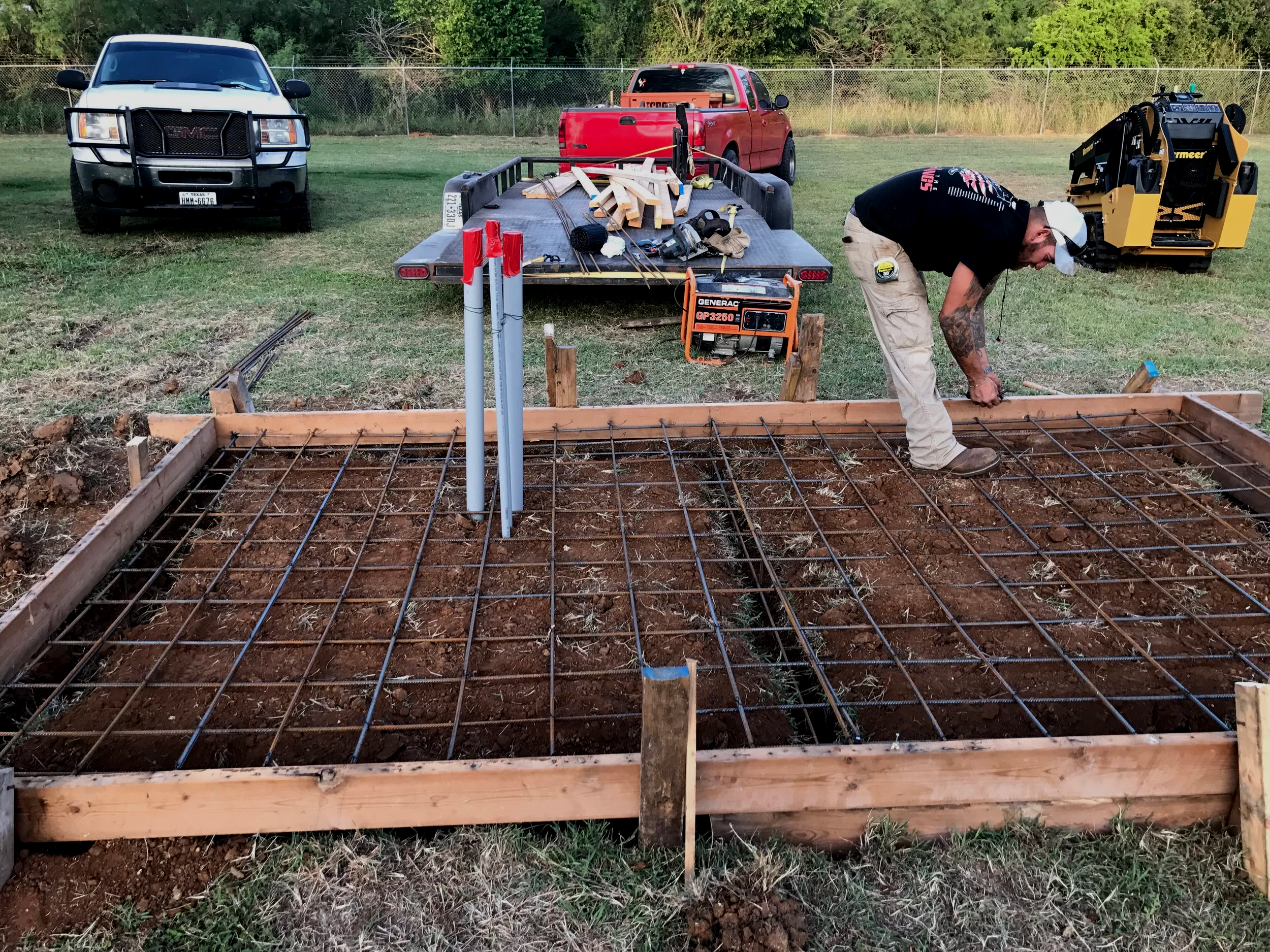 Construction site with a wooden frame foundation, metal rebar, and a worker in a black shirt and beige pants bending over. Two trucks and a trailer loaded with wooden planks and tools are parked on grass, with a fence and trees in the background.