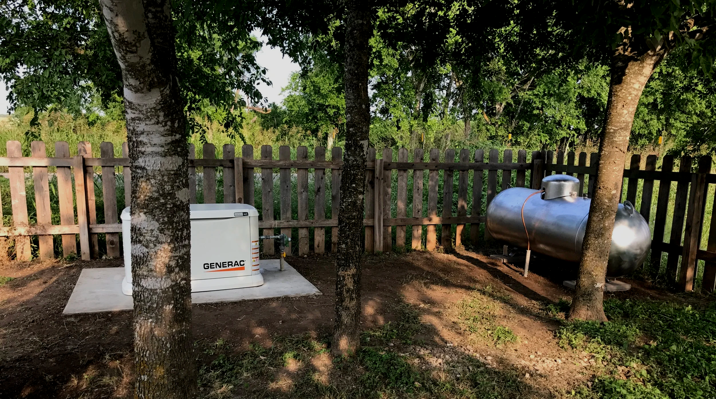 A backyard with a wooden fence, trees, a generator, and a large metal propane tank placed on a concrete pad.