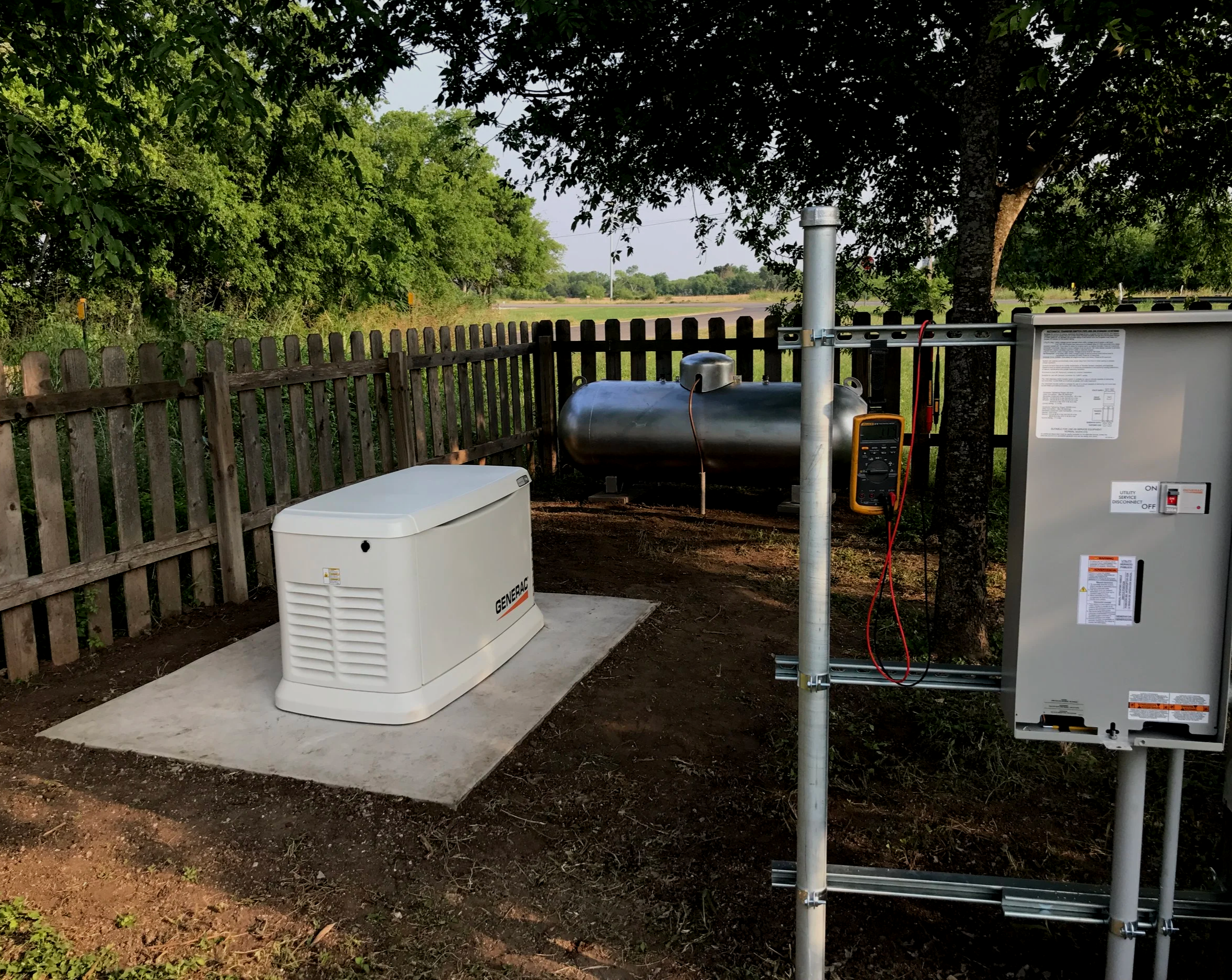 A backyard scene with a wooden fence, trees, and green foliage. In the foreground, there are various electrical equipment, including a generator, a utility disconnect box, and an electrical panel.