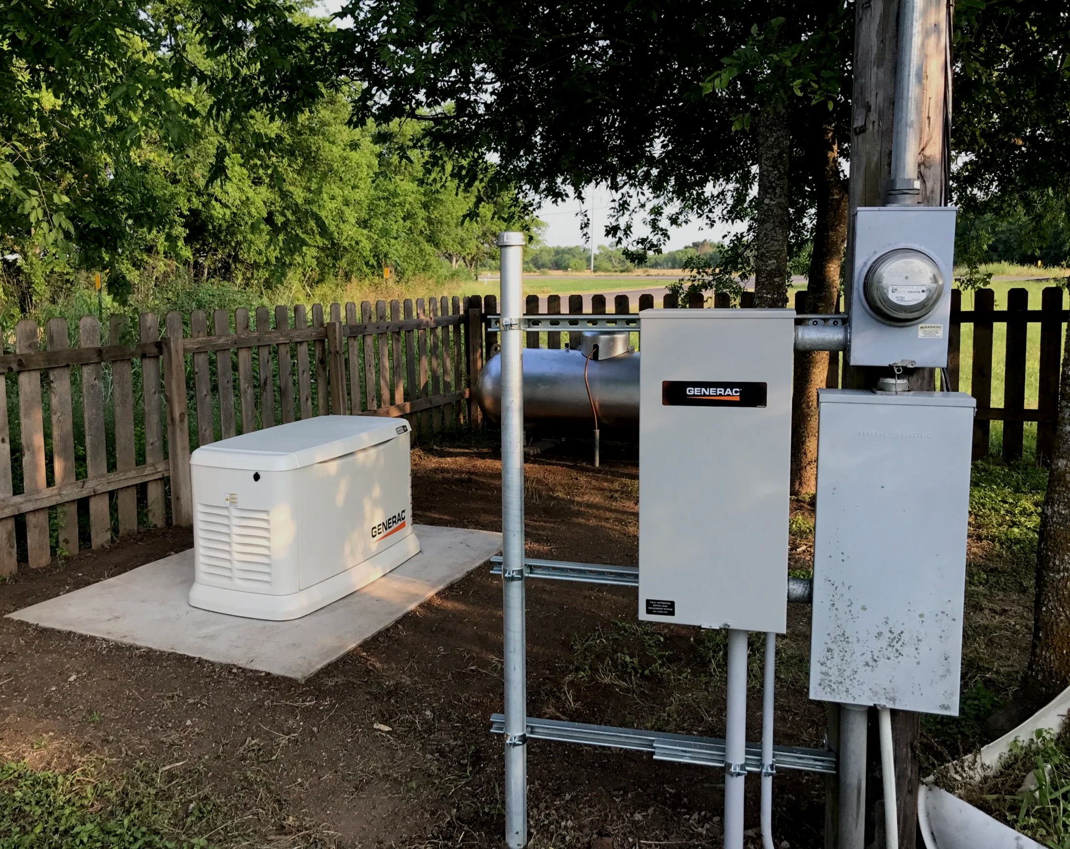 An outdoor scene with electrical equipment, including a generator, electrical boxes, and meters installed on a wooden utility pole. There is a wooden fence, trees, and open land in the background.