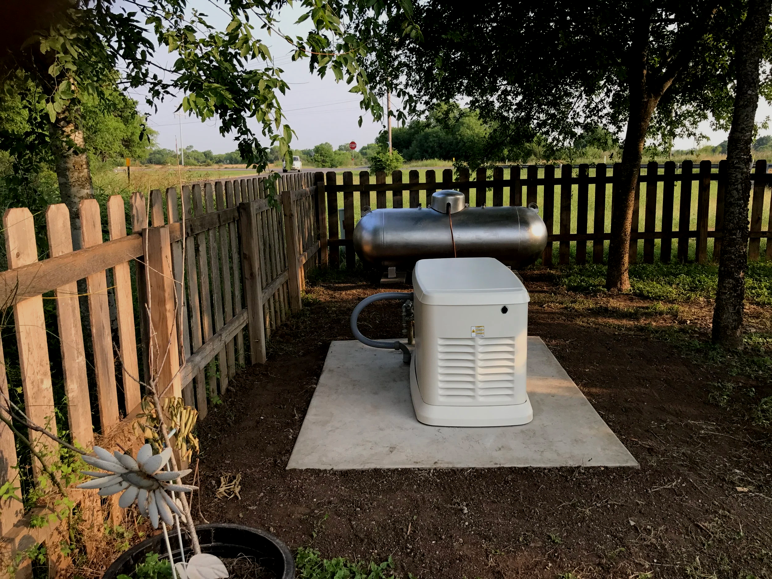 A backyard scene with a wooden fence, trees, and a propane tank on a concrete pad. There is a white appliance or generator connected to a hose, with a small garden or decorative plants nearby. In the background, the road, a car, a stop sign, and open