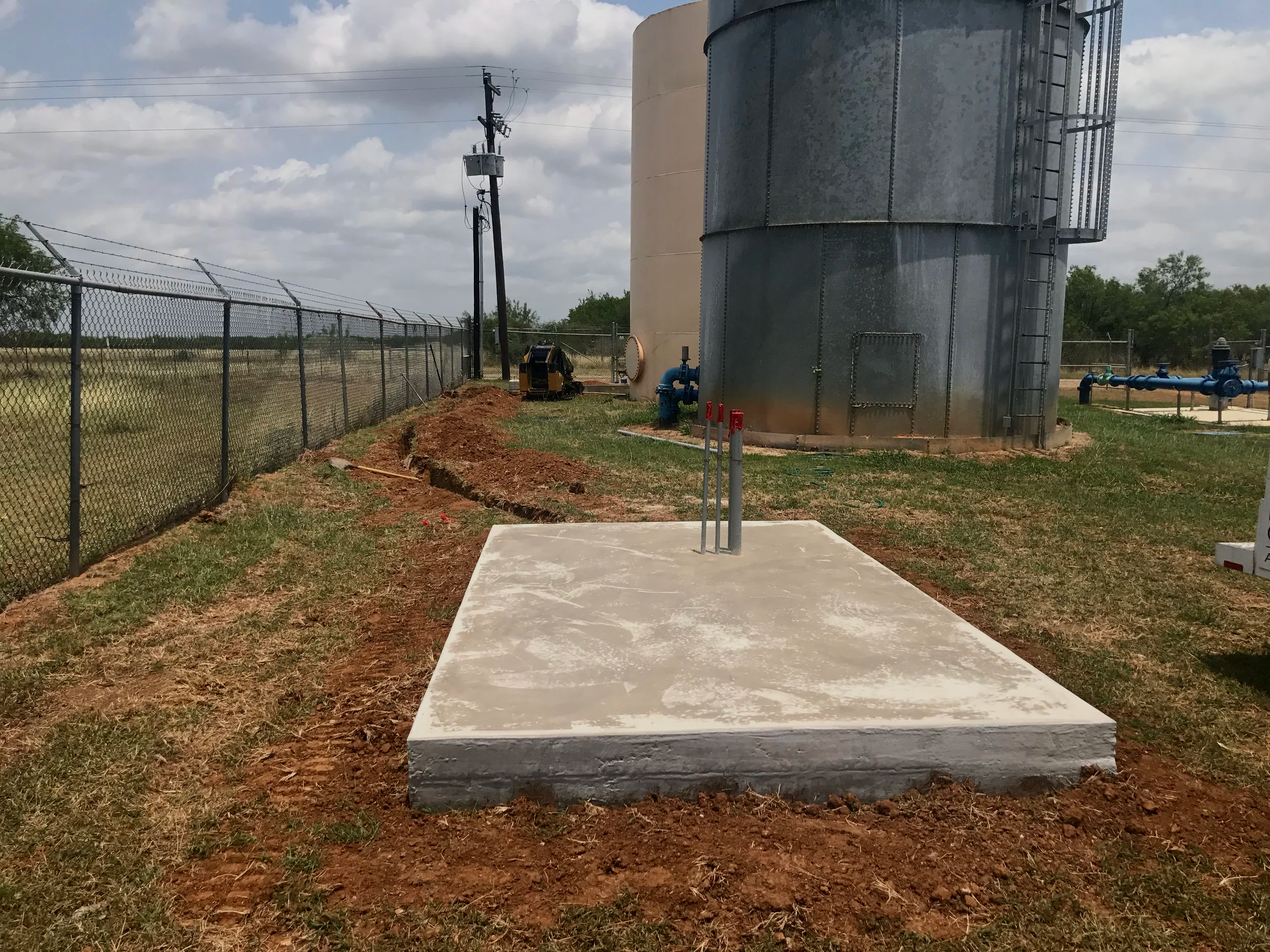 Construction site featuring a large industrial storage tank, a small concrete platform, construction equipment, and a chain-link fence under a partly cloudy sky.