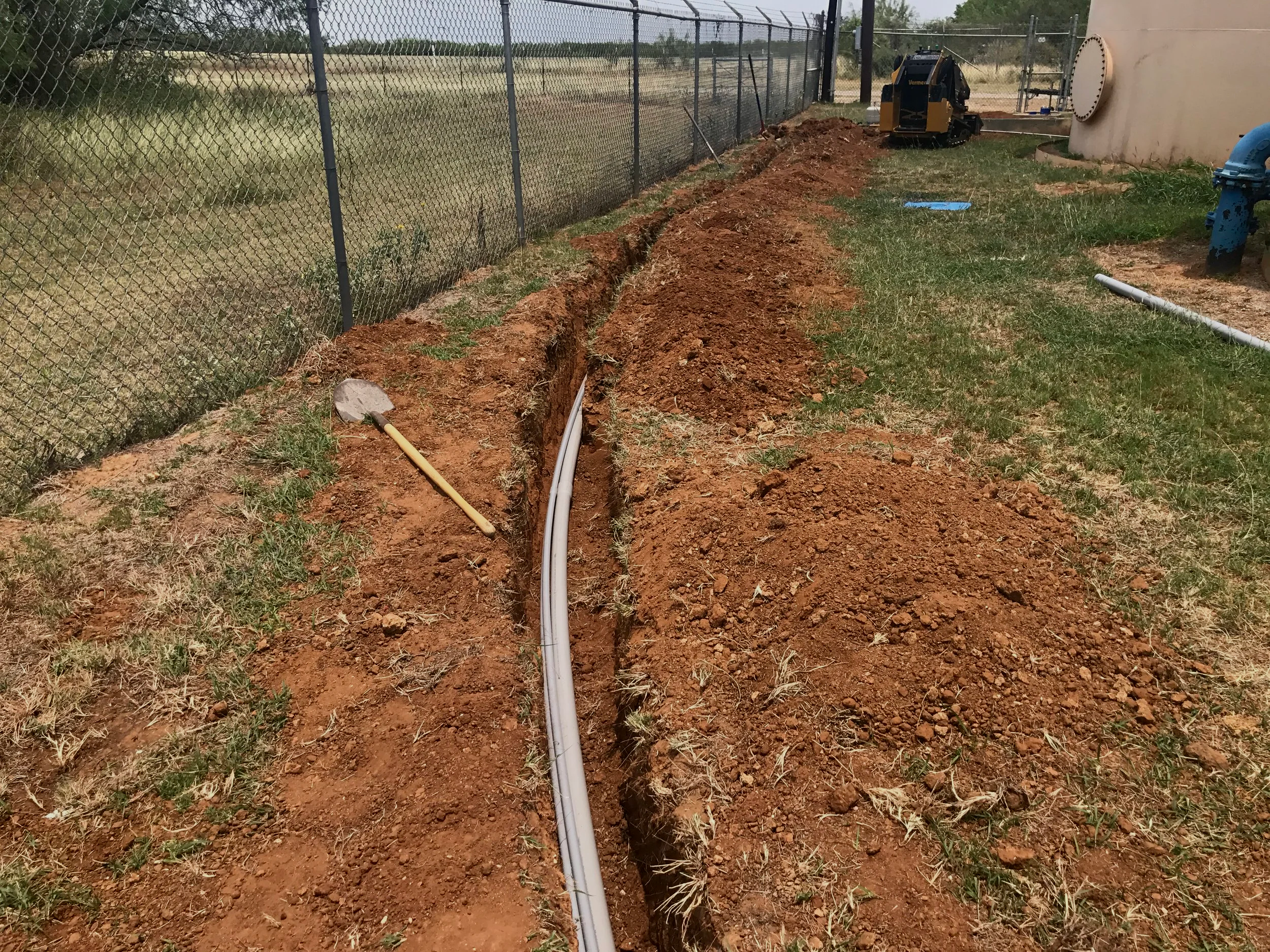A trench dug in the ground with white pipes laid inside, outdoor setting with grass, a chain-link fence, and a small construction vehicle near a white wall.