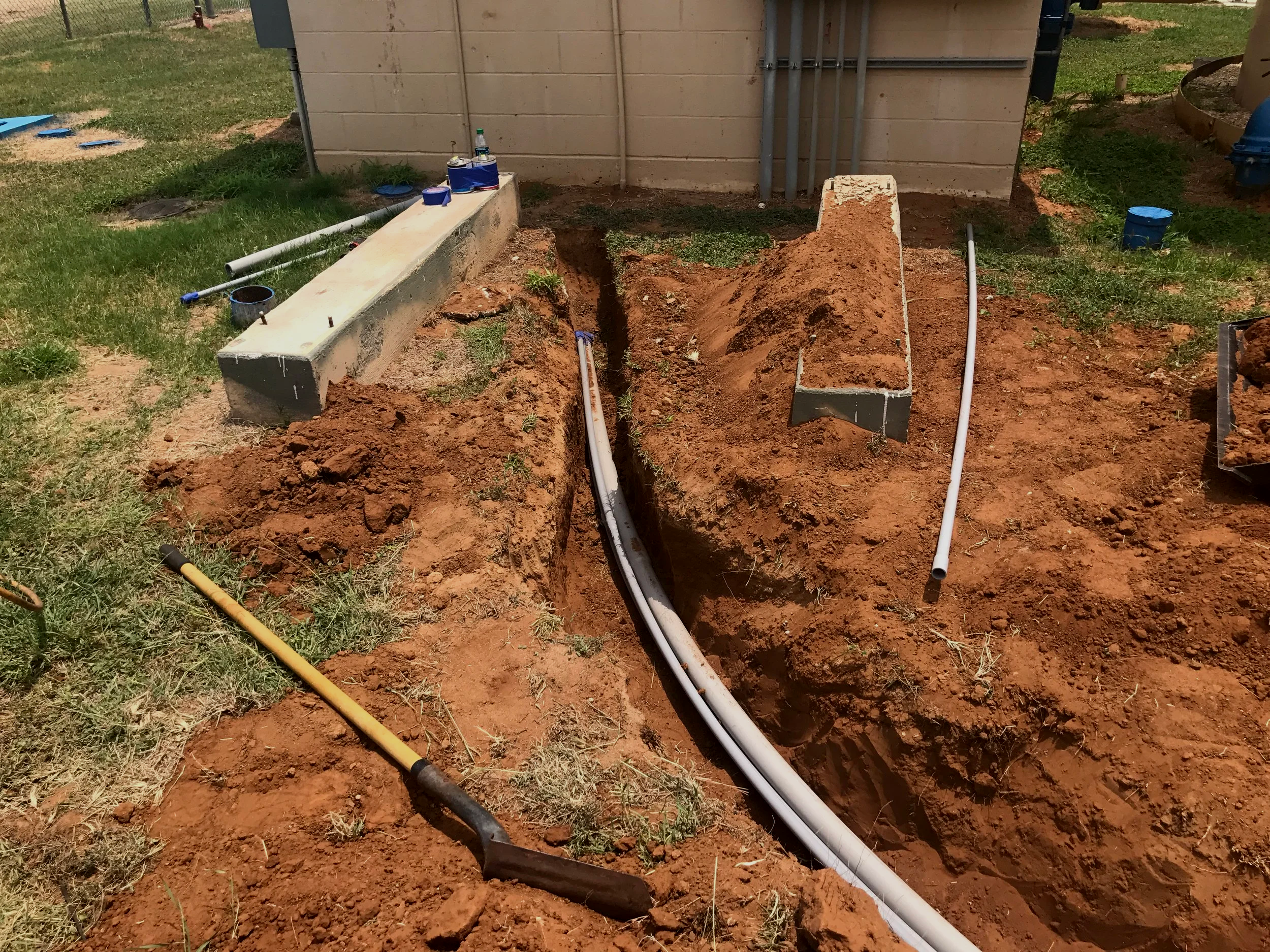 An outdoor construction site with an excavation trench for plumbing pipes, a concrete foundation, construction tools, and building materials nearby.
