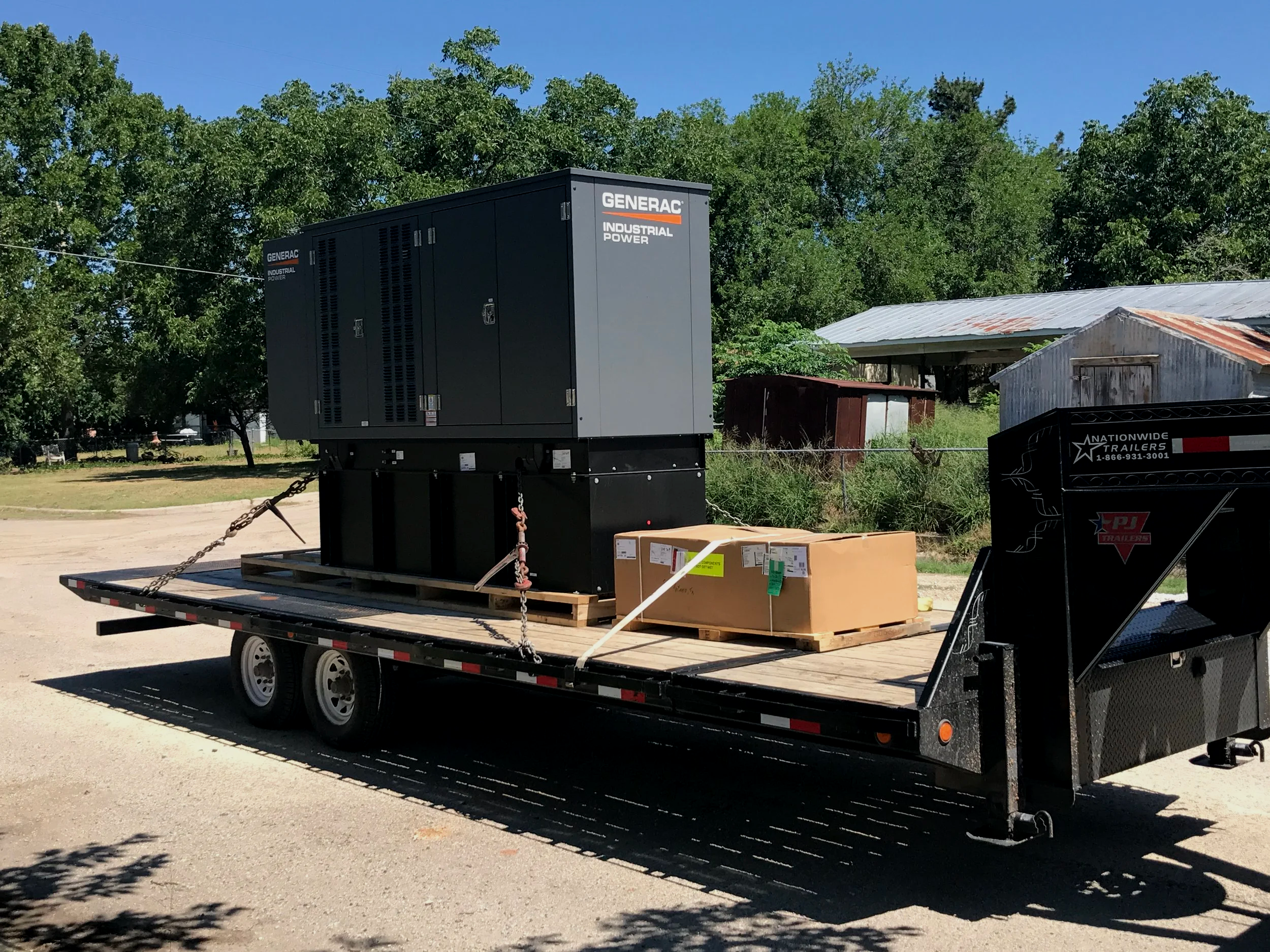 A flatbed trailer transporting a large black Generac industrial power generator with trees and a building in the background.