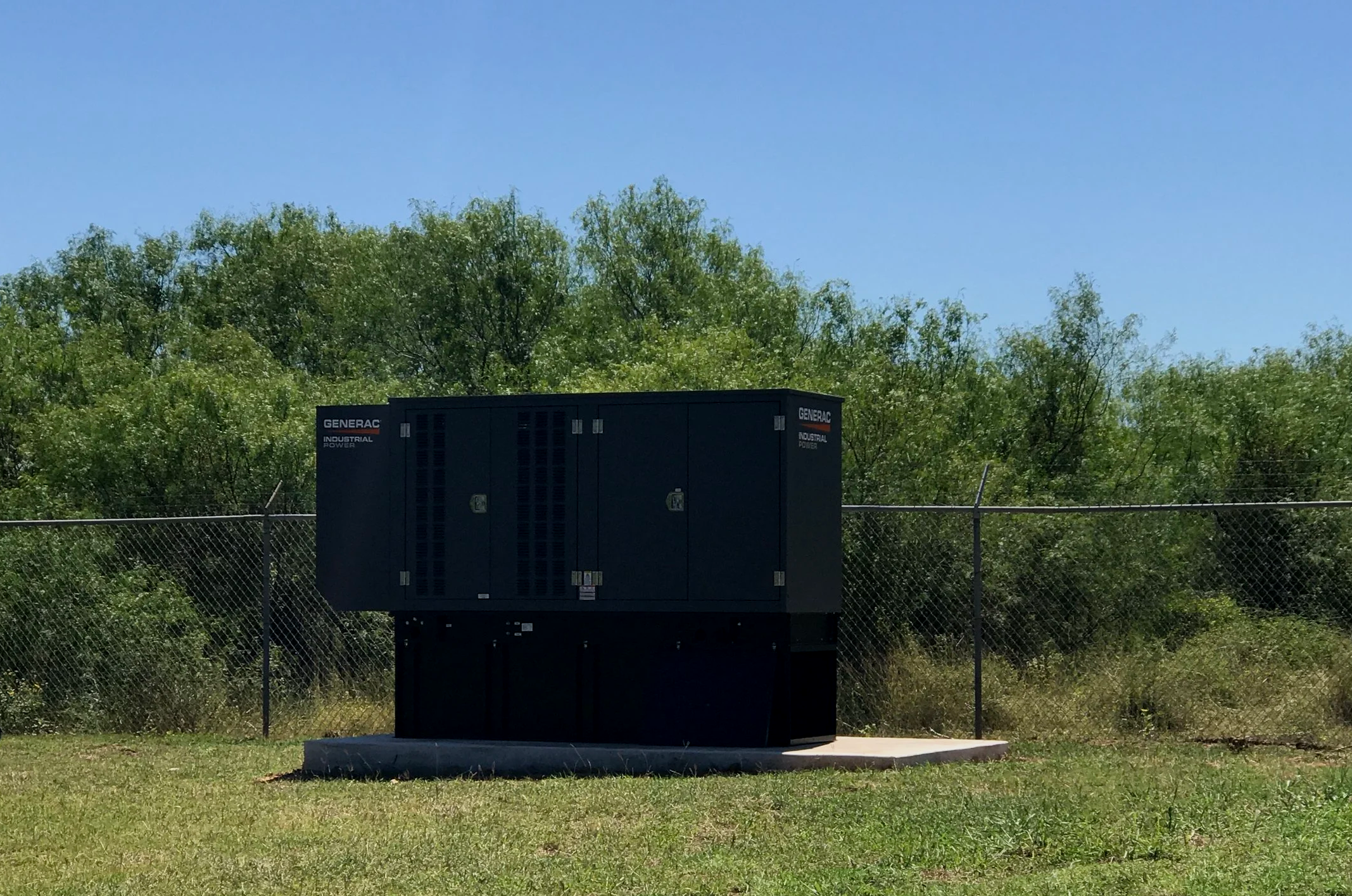 An electrical generator labeled 'Generac Industrial Power' is situated outdoors on a concrete slab, with a chain-link fence in the background and green trees and a clear blue sky overhead.