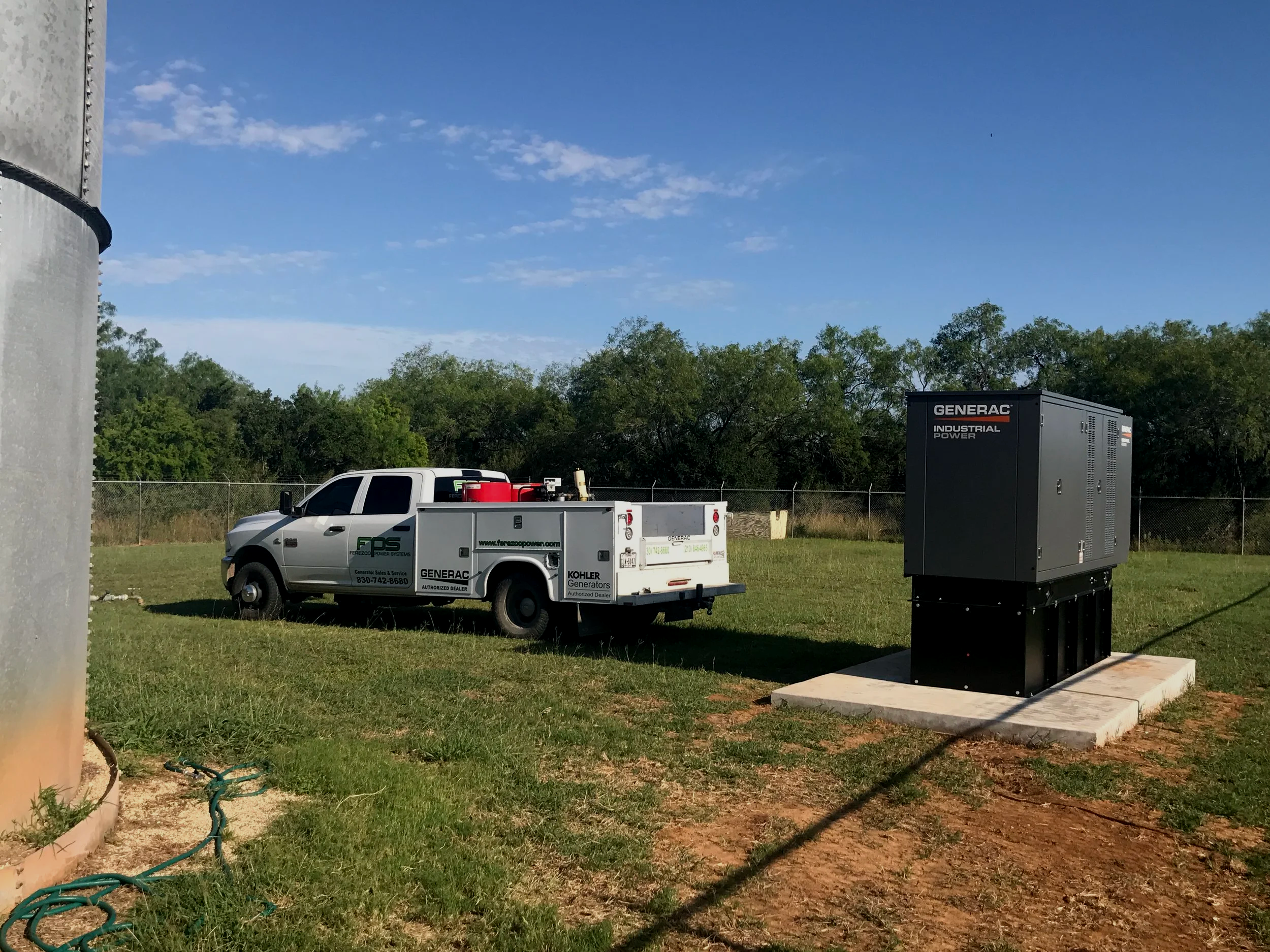 A utility yard with a large generator, a service truck, and a smaller generator on a grassy area with trees and blue sky in the background.