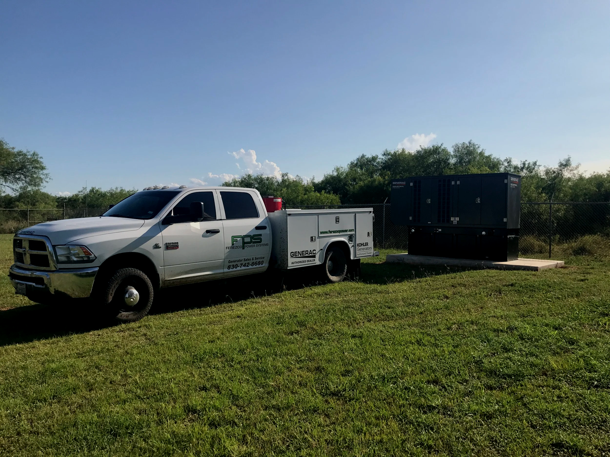A white utility truck with green decals and text, parked on a grassy area next to a fenced landscape with trees, under a clear blue sky with some clouds and the sun.