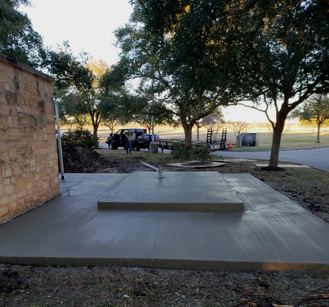 Freshly poured concrete sidewalk next to a stone building, with trees and a street in the background, during sunset.