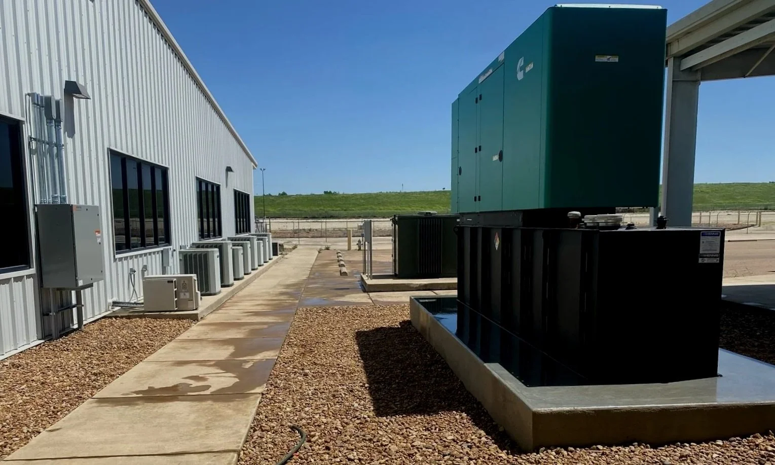 Outdoor industrial area with a metal building, multiple air conditioning units, and large green and black electrical or generator units on concrete slabs, under a clear blue sky.