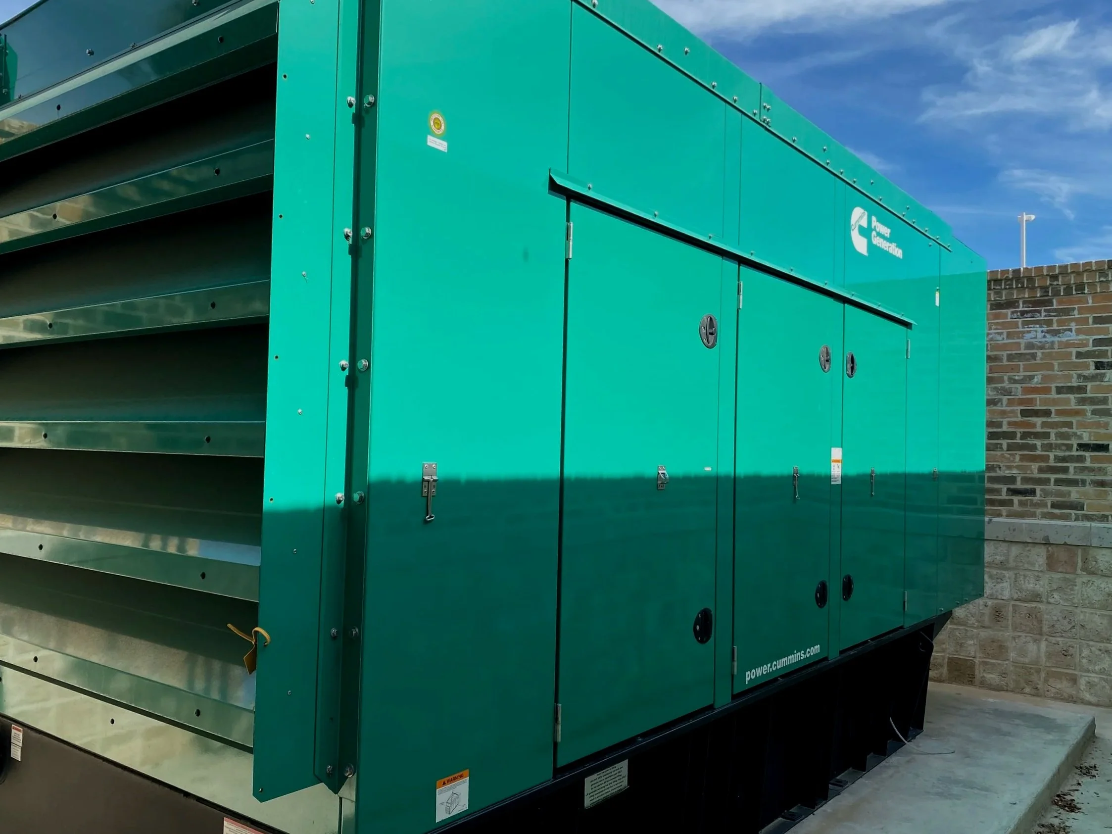 Large teal-colored industrial generator with vents and locks, situated outdoors against a brick wall with a flagpole in the background, under a blue sky with some clouds.