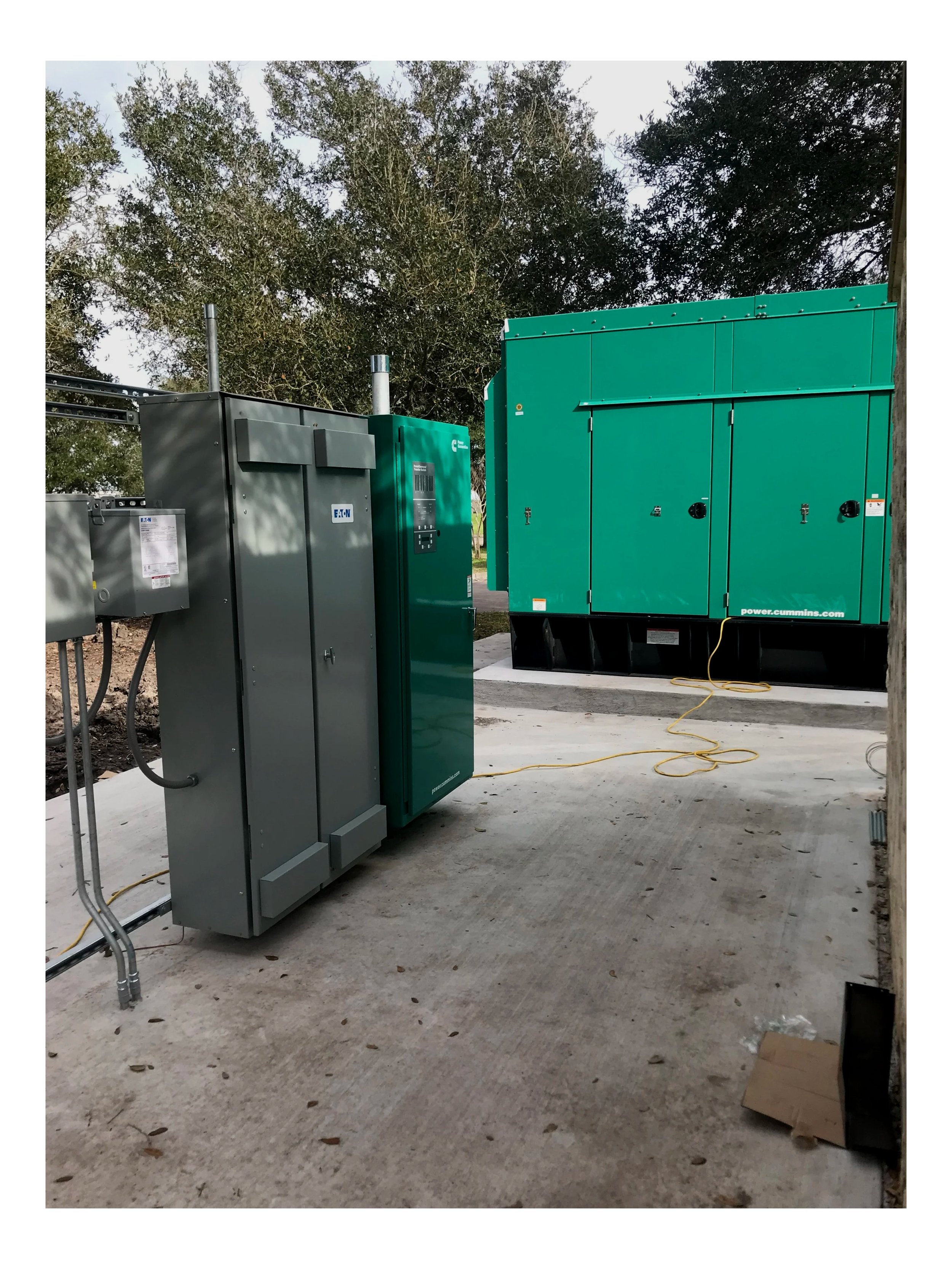 Numerous large green and gray electrical equipment boxes and a generator at an outdoor utility site with trees in the background and a concrete floor.