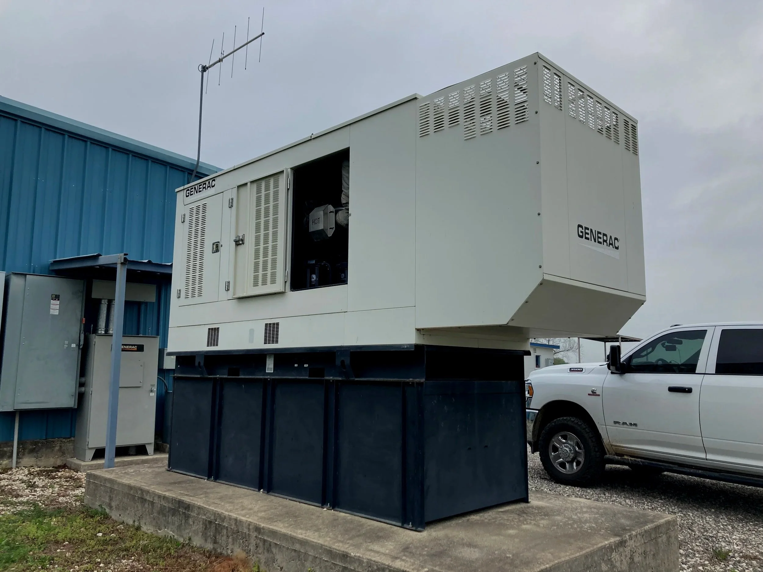 Industrial generator unit labeled 'Generac' installed outdoors on a concrete platform with a white pickup truck nearby and a blue building in the background.