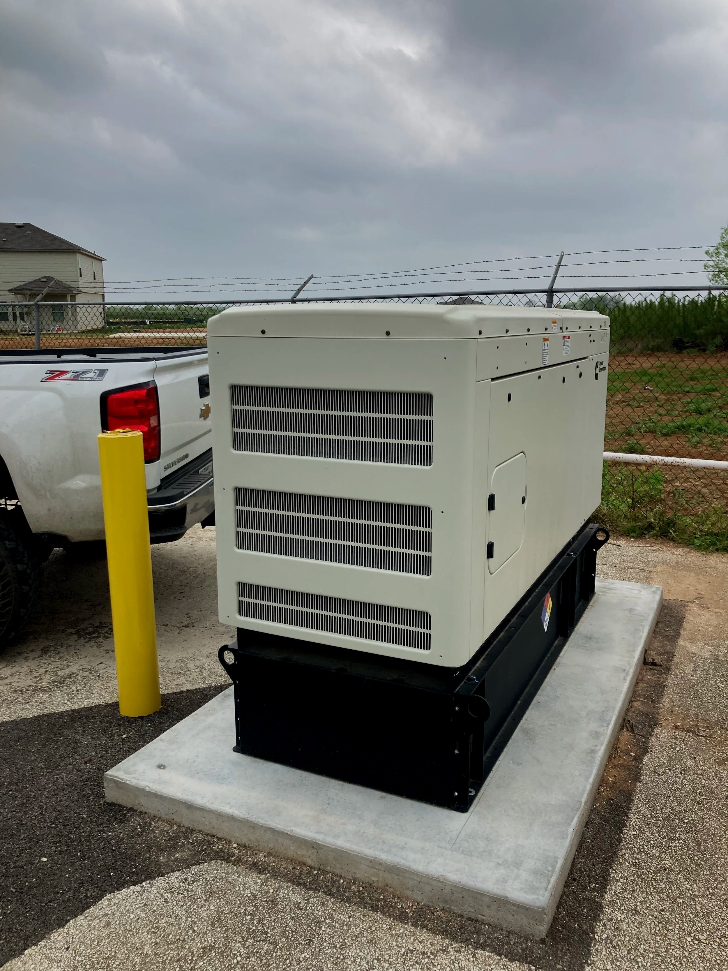 A large white generator installed on a concrete pad in an outdoor setting, with a black base, surrounded by a chain-link fence, and a pickup truck parked nearby. Overcast sky in the background.