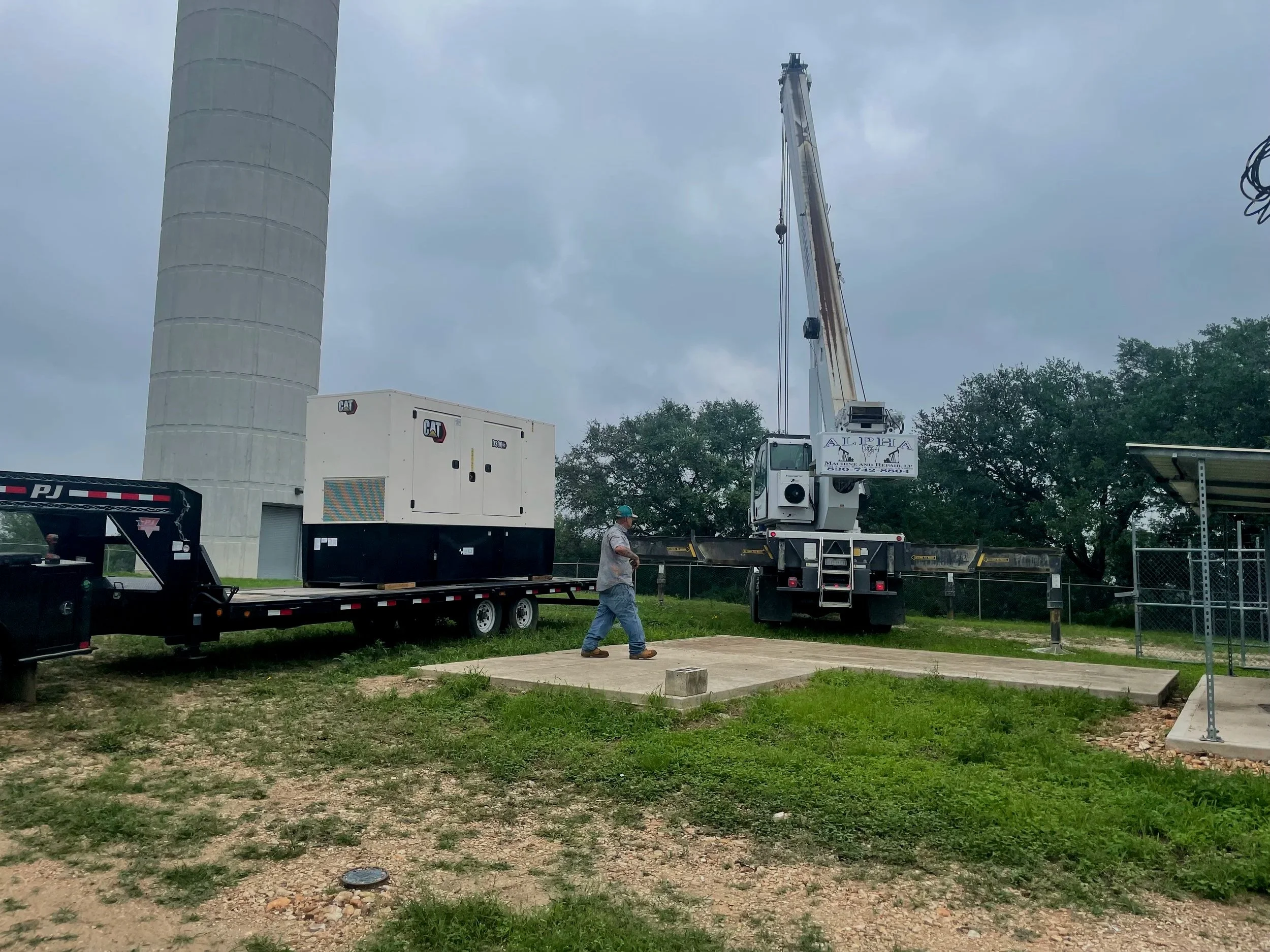A construction scene showing a large crane lifting equipment, a man walking on a concrete slab, and construction equipment with power generators outdoors under cloudy skies.