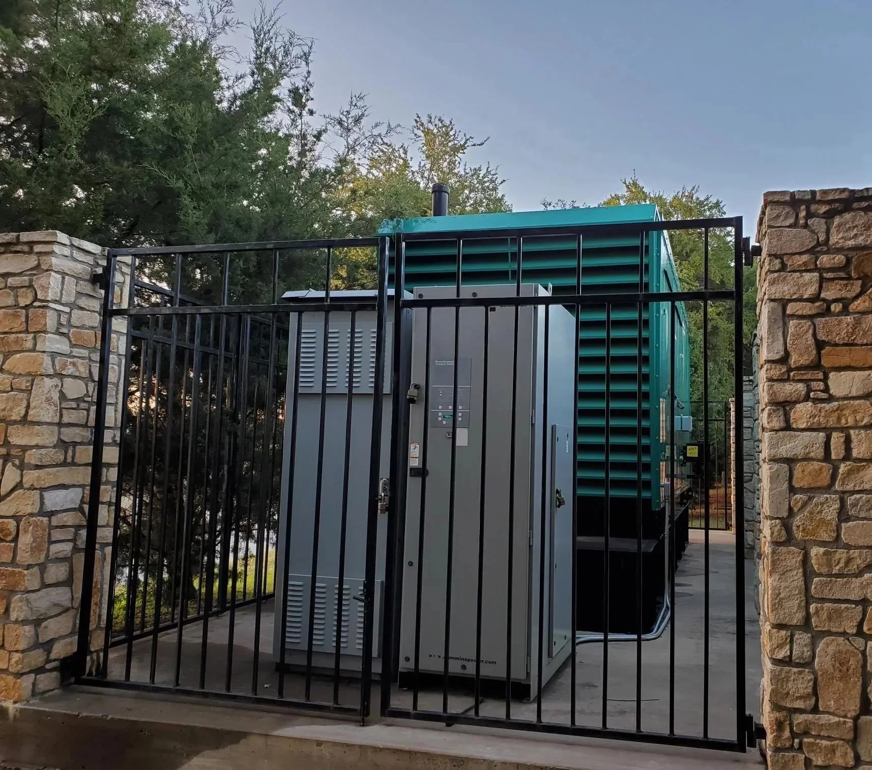 A security gate enclosing an electrical transformer unit, with stone pillars on either side and a black metal fence.