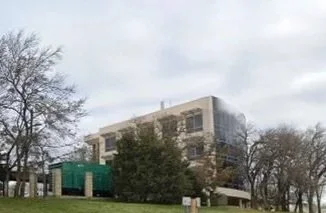 A multi-story building with trees and a park in the foreground under a cloudy sky.