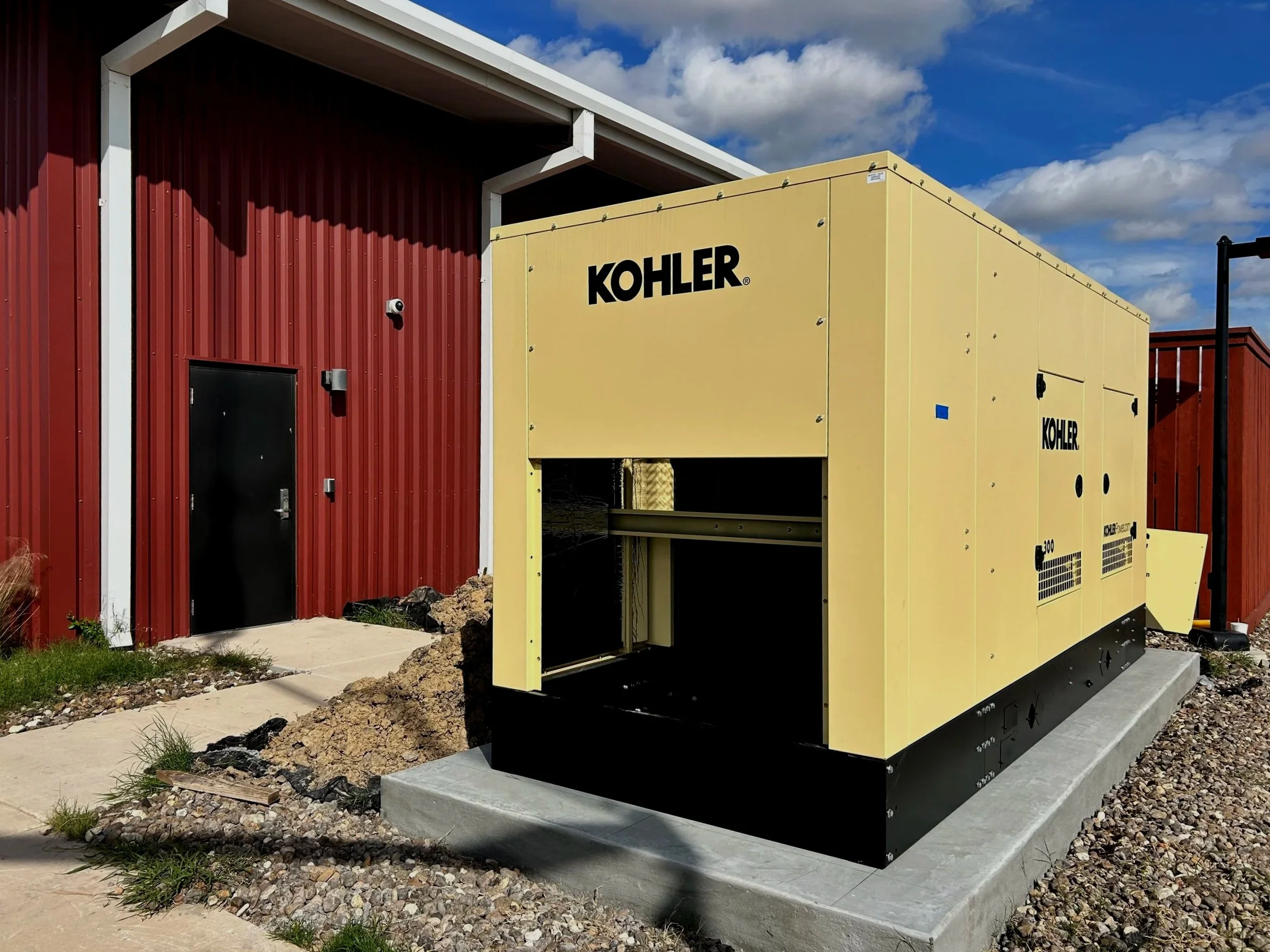Yellow KOHLER generator installed outdoors in a residential setting on a concrete pad, next to a red building with a small security camera mounted on the wall.