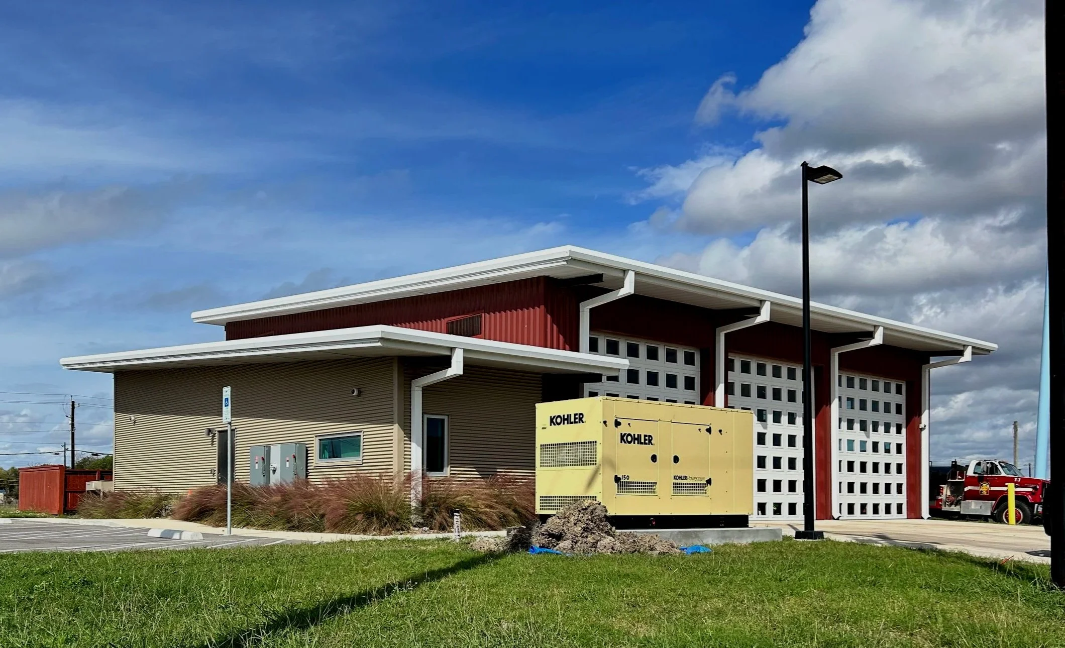 A modern building with beige and red exterior walls, a white roof, and multiple small windows. In front, there is a yellow Kohler generator, a parking lot with lines and a handicap parking sign, and some grass and landscaping. A blue sky with clouds 