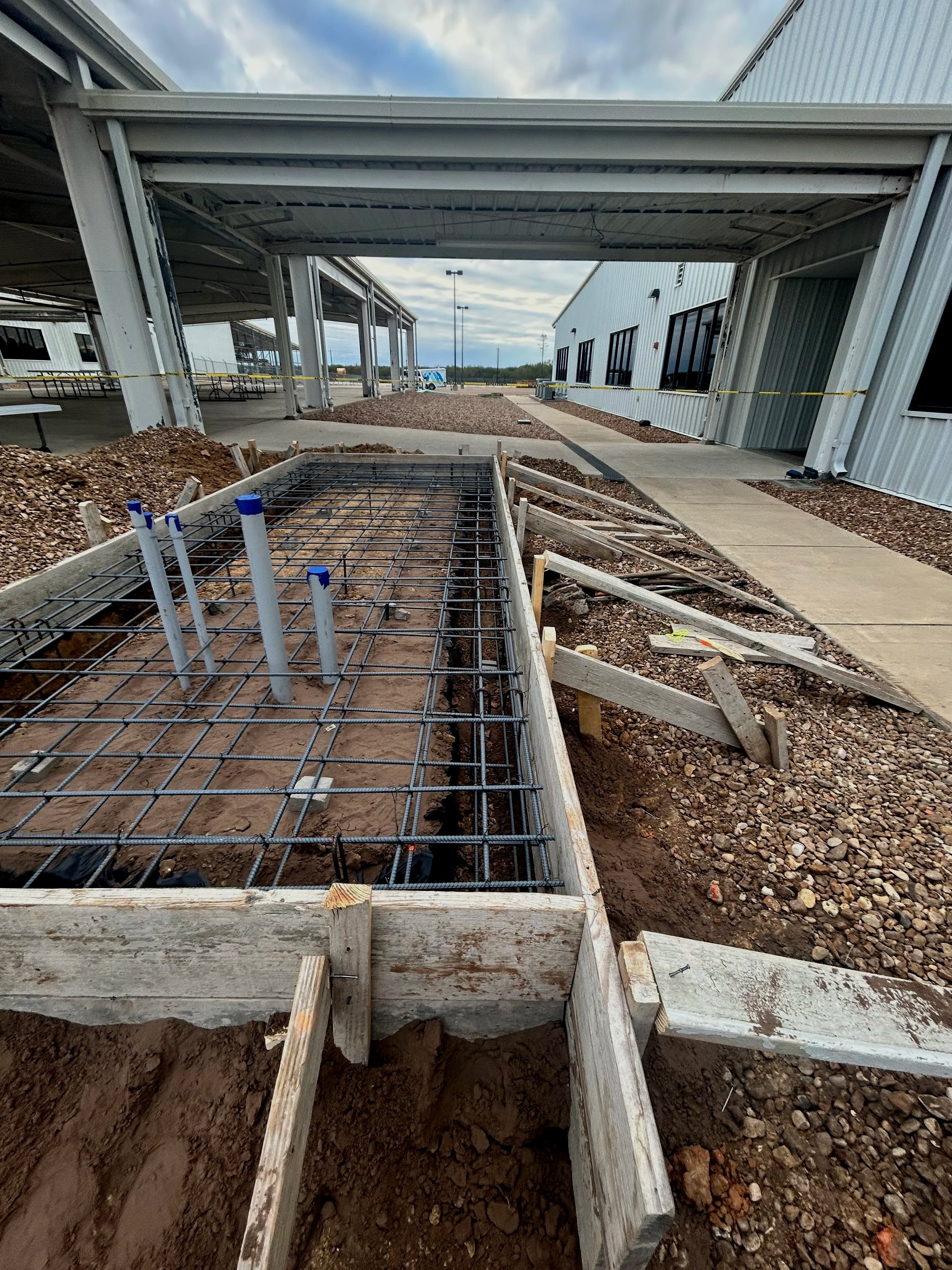 Construction site showing a concrete pathway, wooden forms, and metal rebar framework for pouring a concrete slab, with gravel and dirt surrounding the area, and covered buildings in the background.