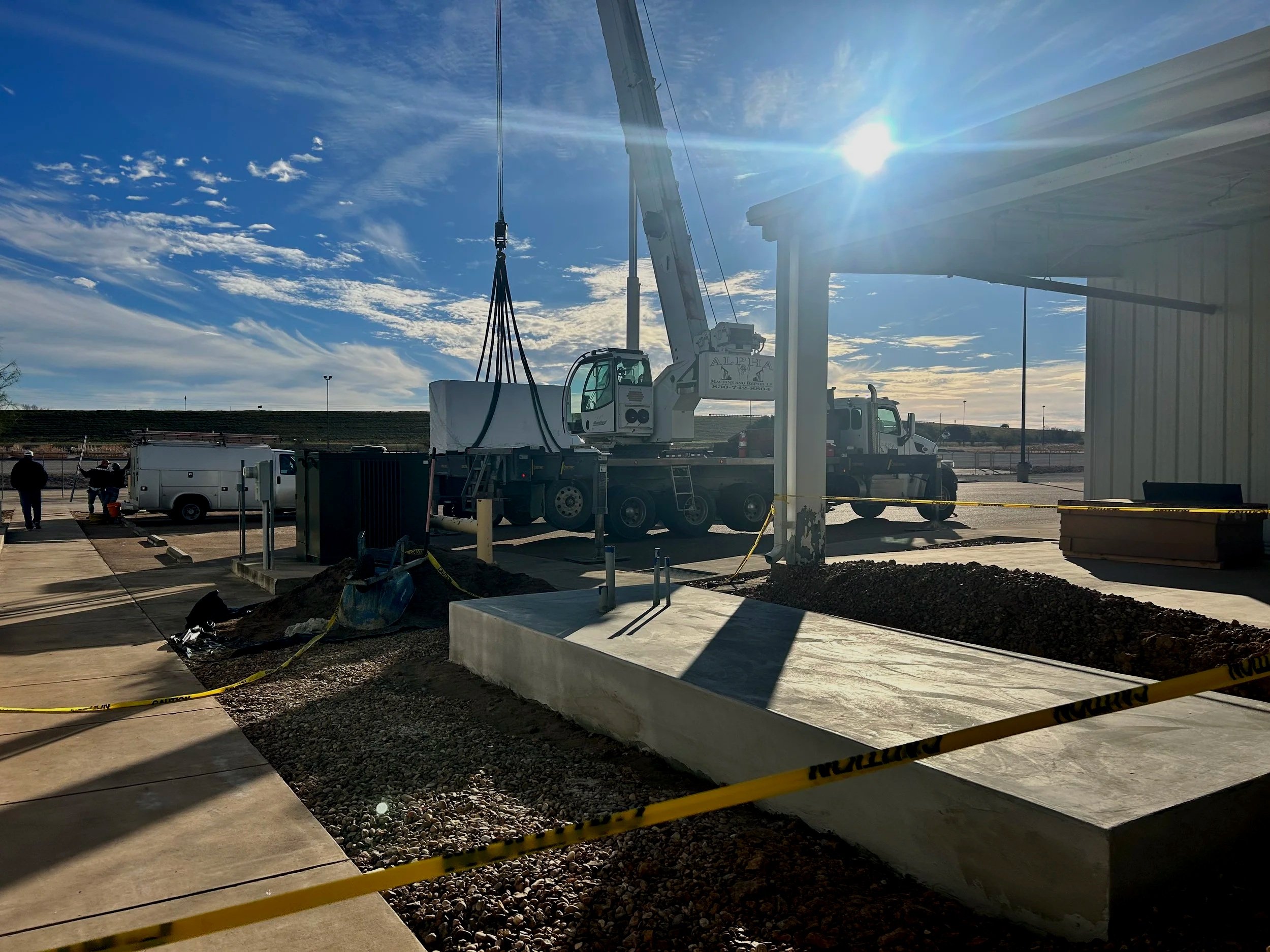 Construction site with a crane lifting materials, workers, and caution tape in the foreground under a sky with sunlight and clouds.